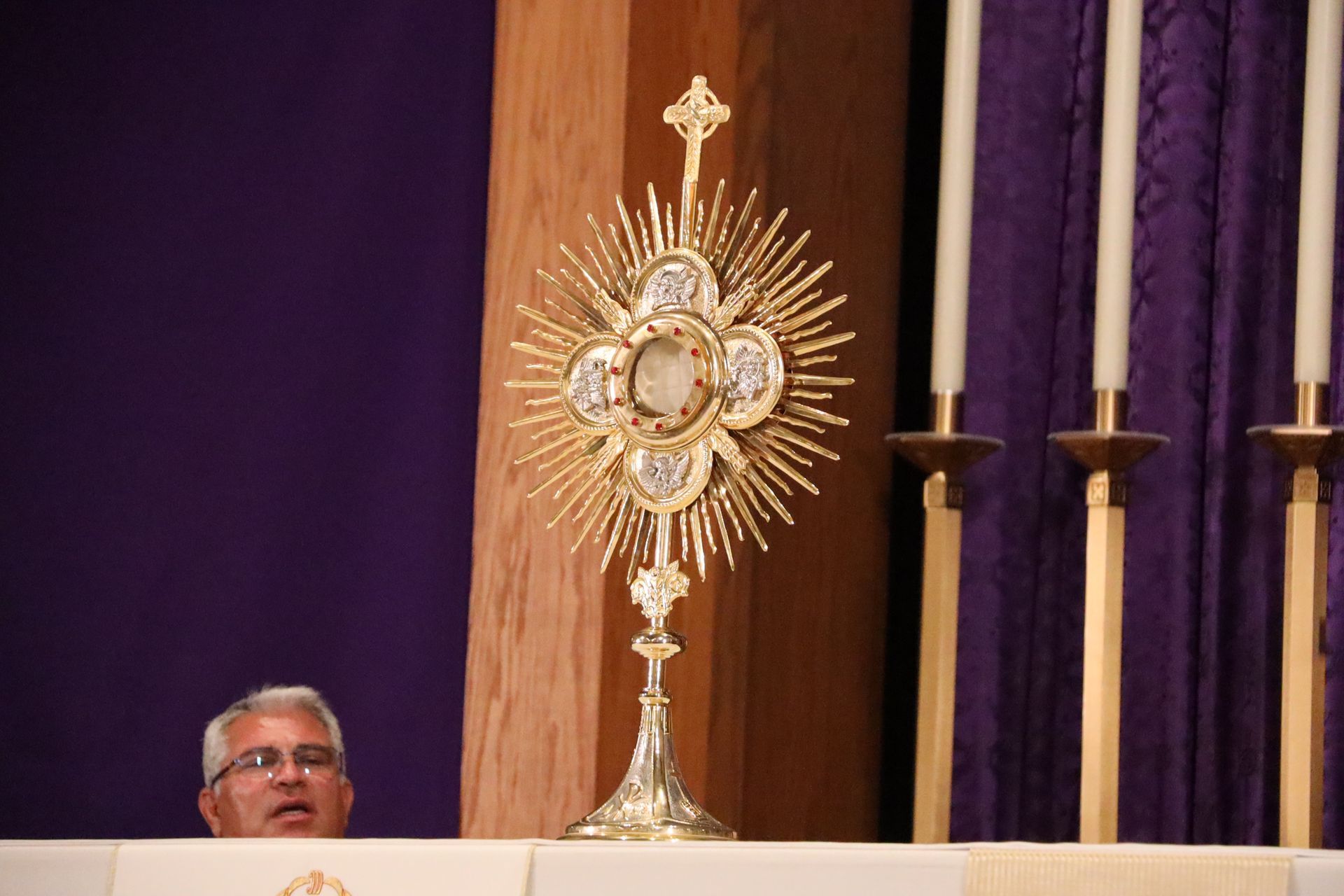 Monstrance on an altar before purple drapes, with a priest partially visible behind it