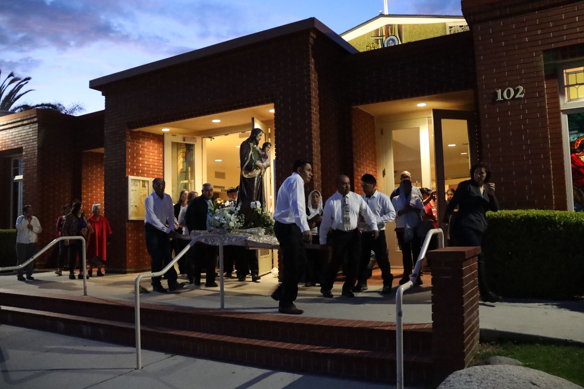 People gathered outside a brick building at dusk, standing near tables by the entrance.