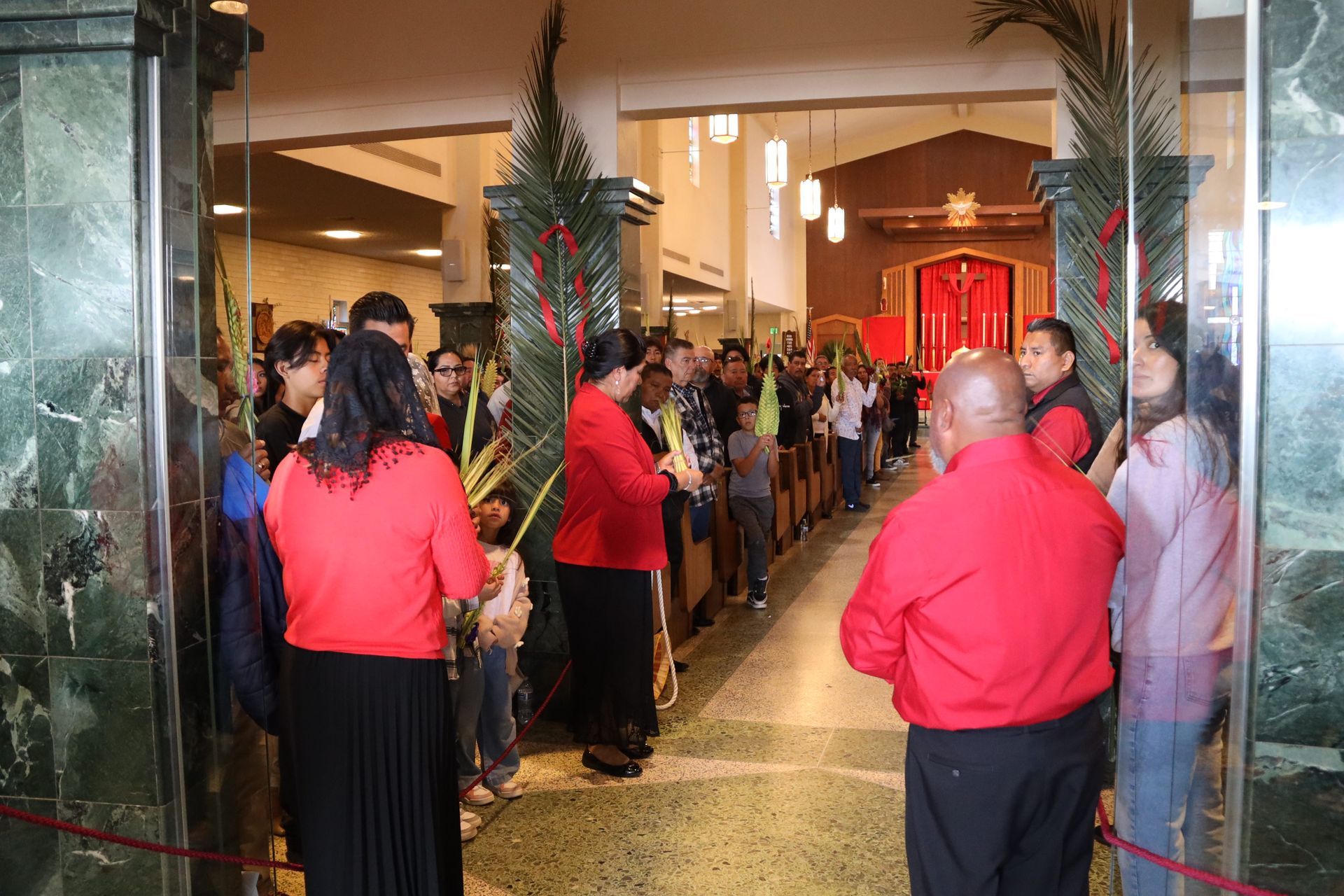 People gathered in a lobby entrance with red-shirted staff standing by glass pillars.