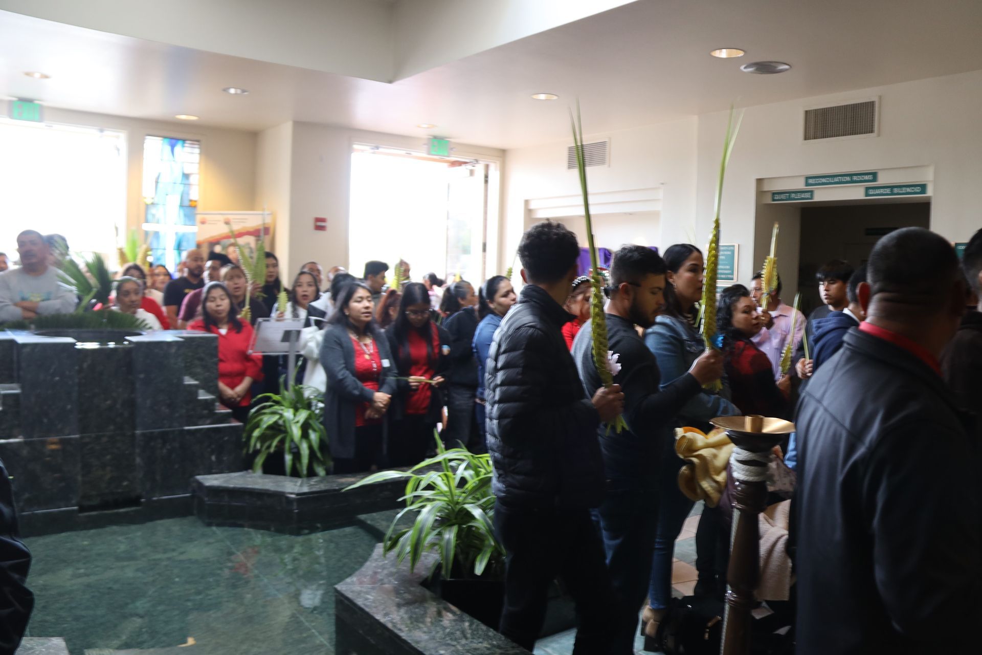 Crowded lobby with people waiting in line near a counter, potted plants, and a bright entrance.