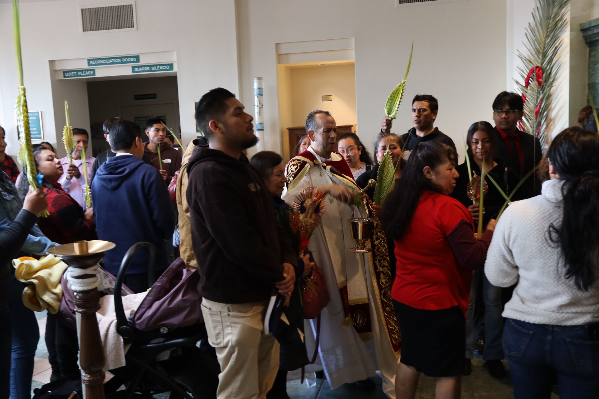 People gathered indoors in a lively crowd, some holding palm branches and flowers during a ceremony.