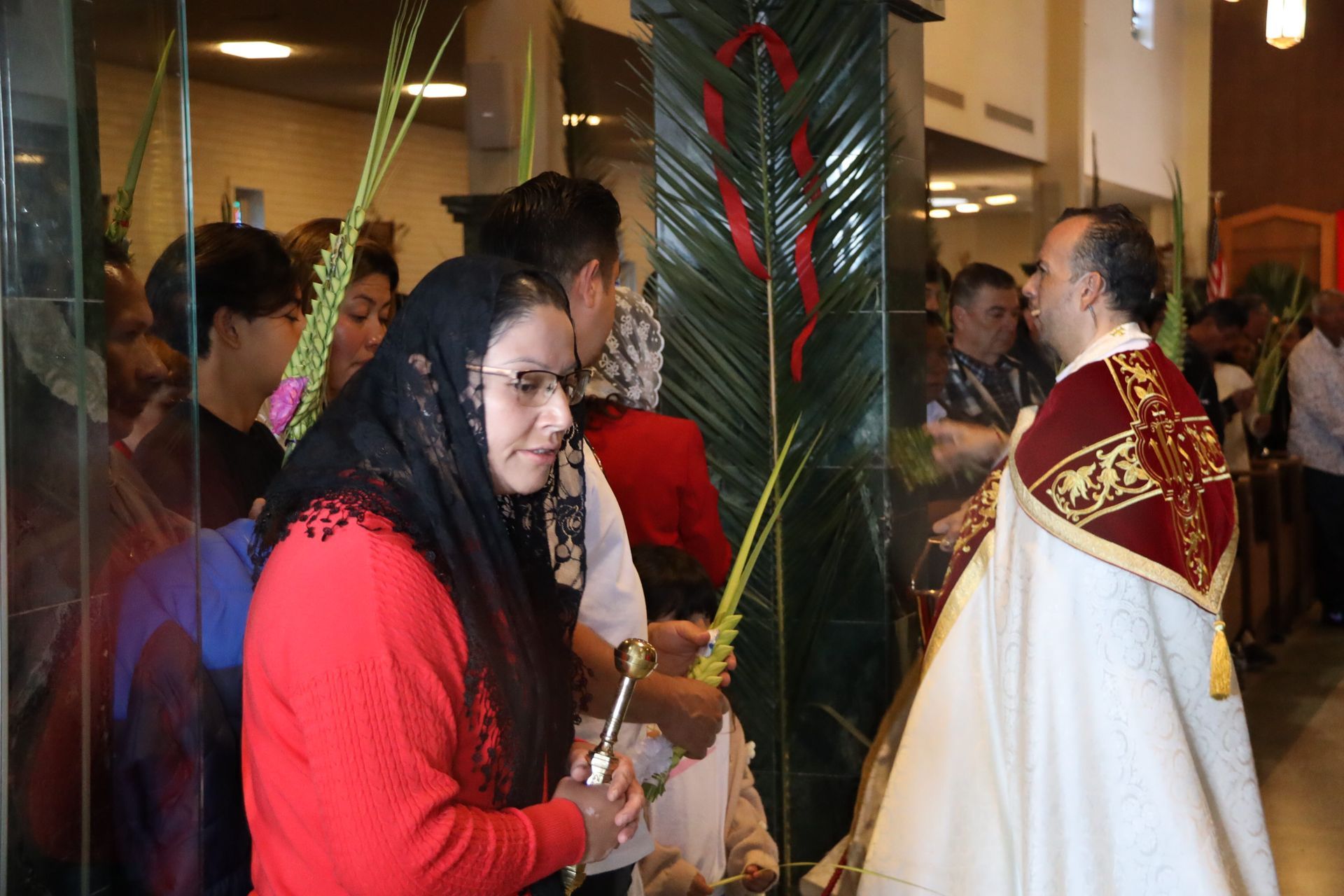People gathered indoors during a religious ceremony, with a priest in a white robe facing the congregation.