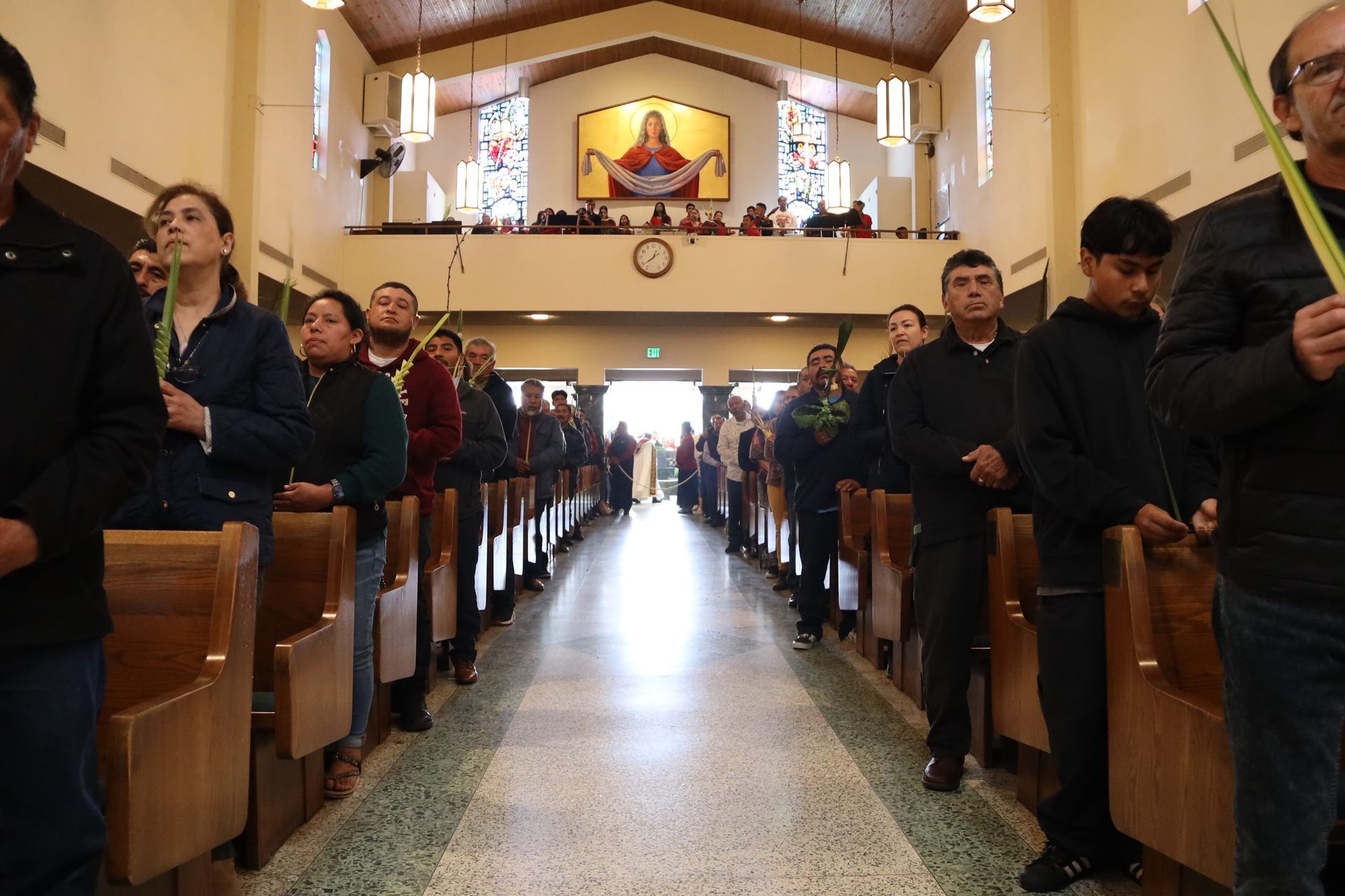 People standing in church pews during a service, with a bright altar at the far end.
