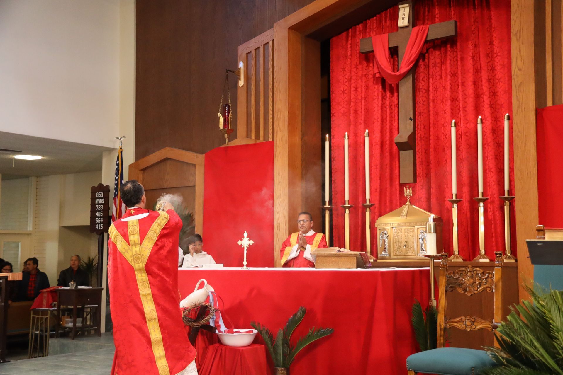 Church altar with red drapery and candles, two clergy in red vestments, one speaking at the lectern.