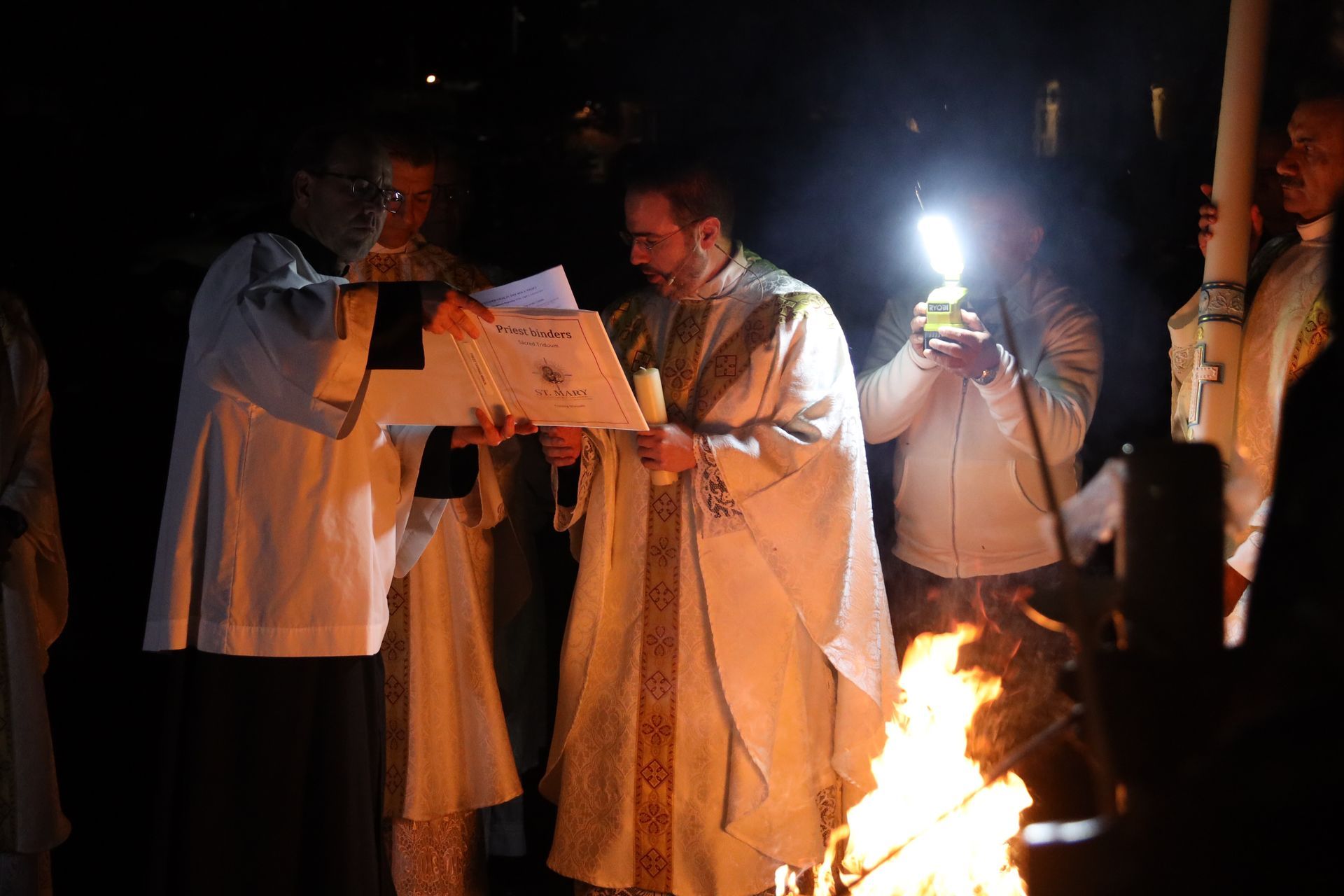People gathered at night around a fire, reading from papers in a dimly lit scene.
