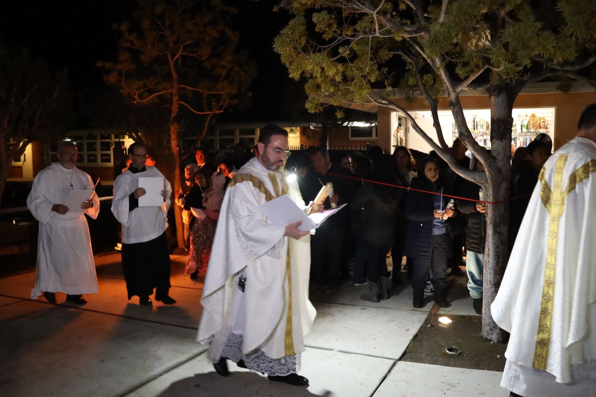 Clergy in white robes conduct an outdoor nighttime ceremony with onlookers gathered around.