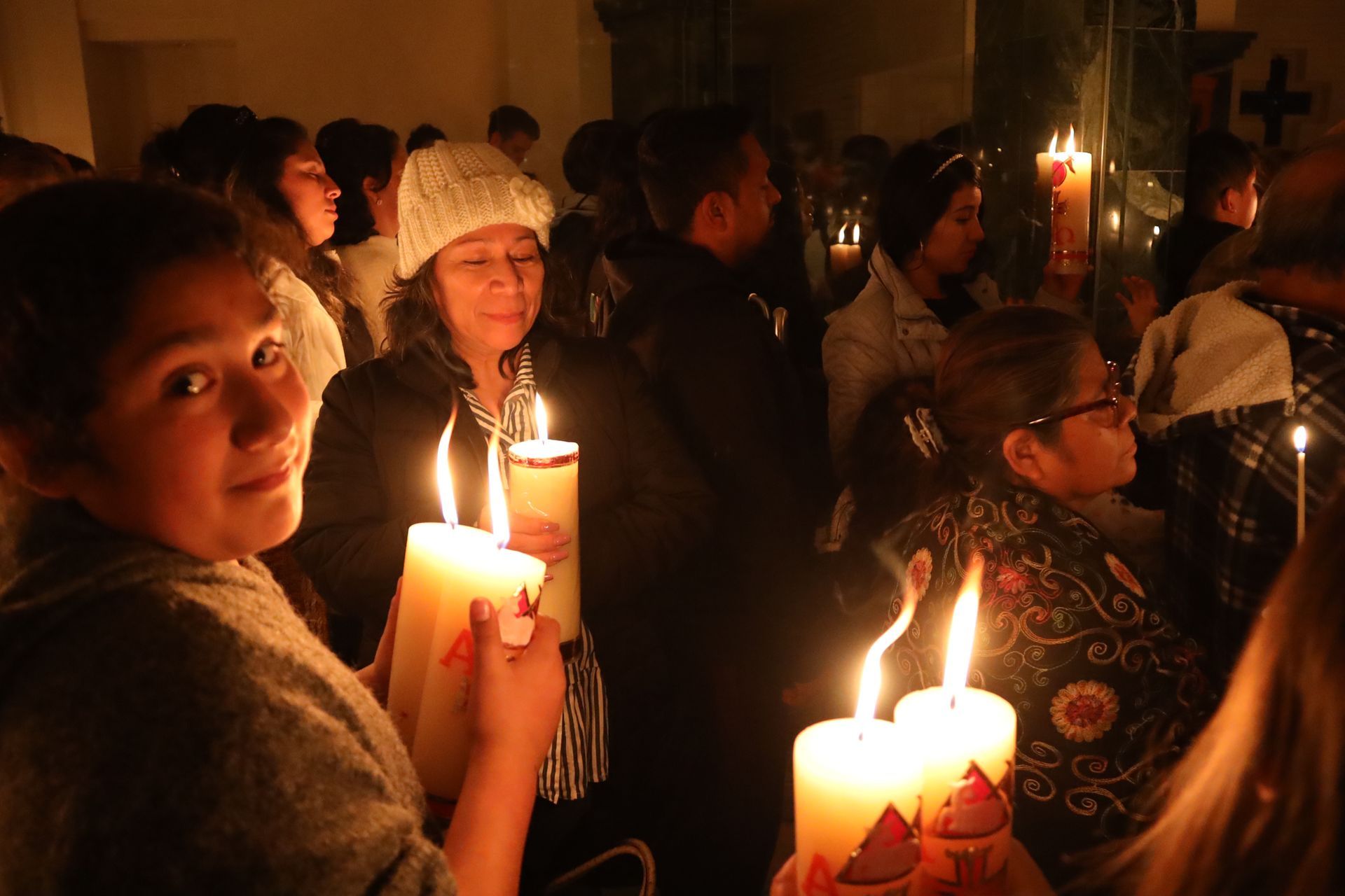 People holding lit candles in a dim indoor gathering, with warm candlelight illuminating their faces.