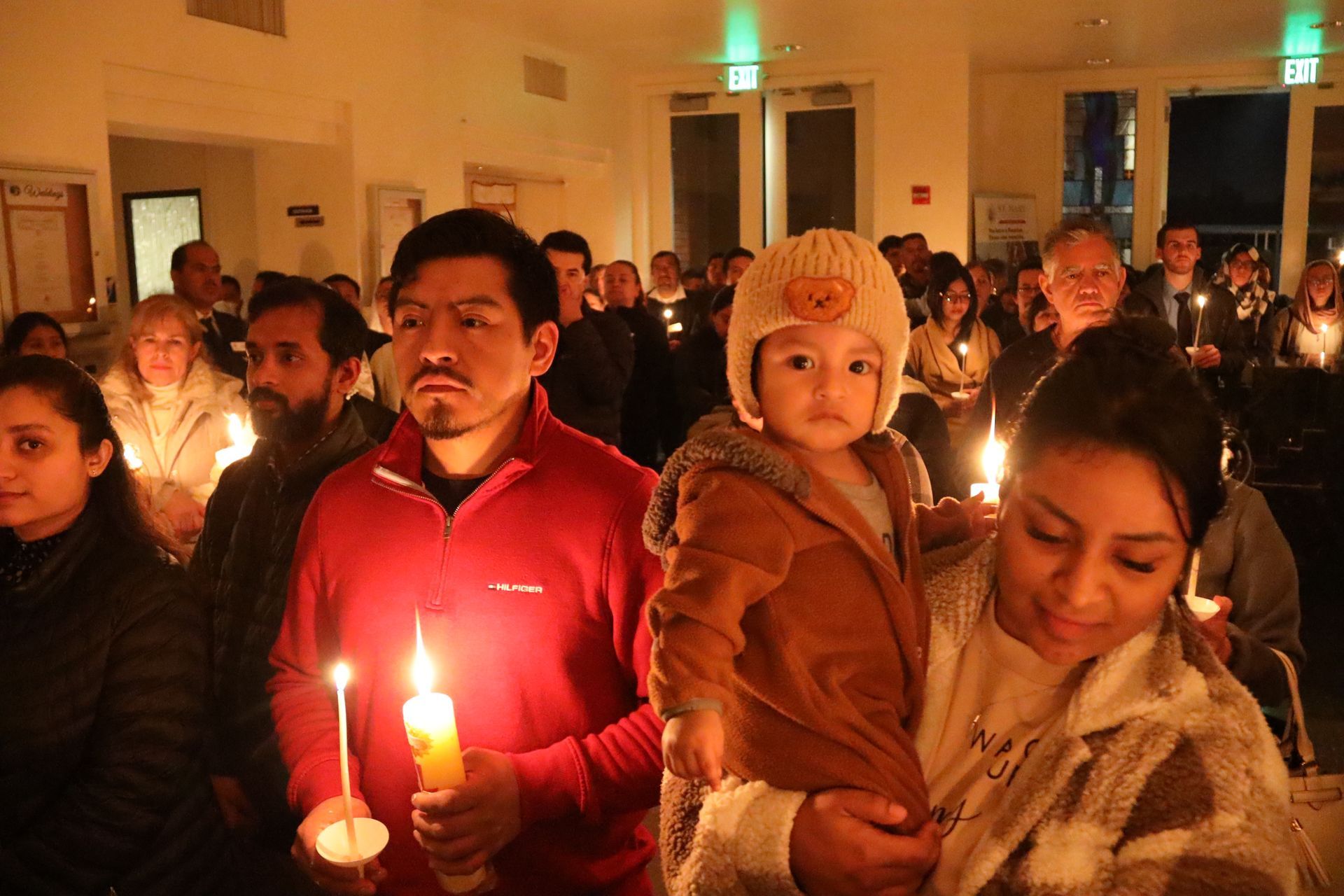 People holding lit candles at an indoor vigil, with warm light and solemn expressions.