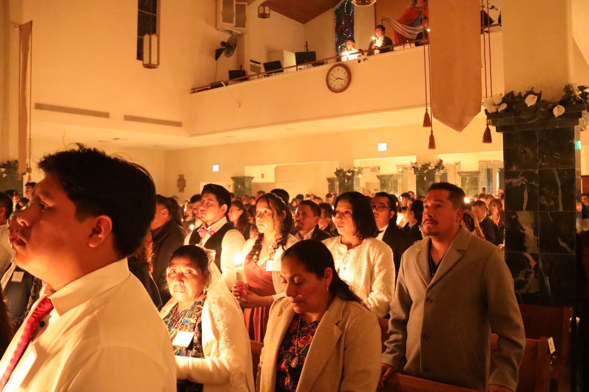 People standing in a warmly lit church or hall, holding candles during a service.