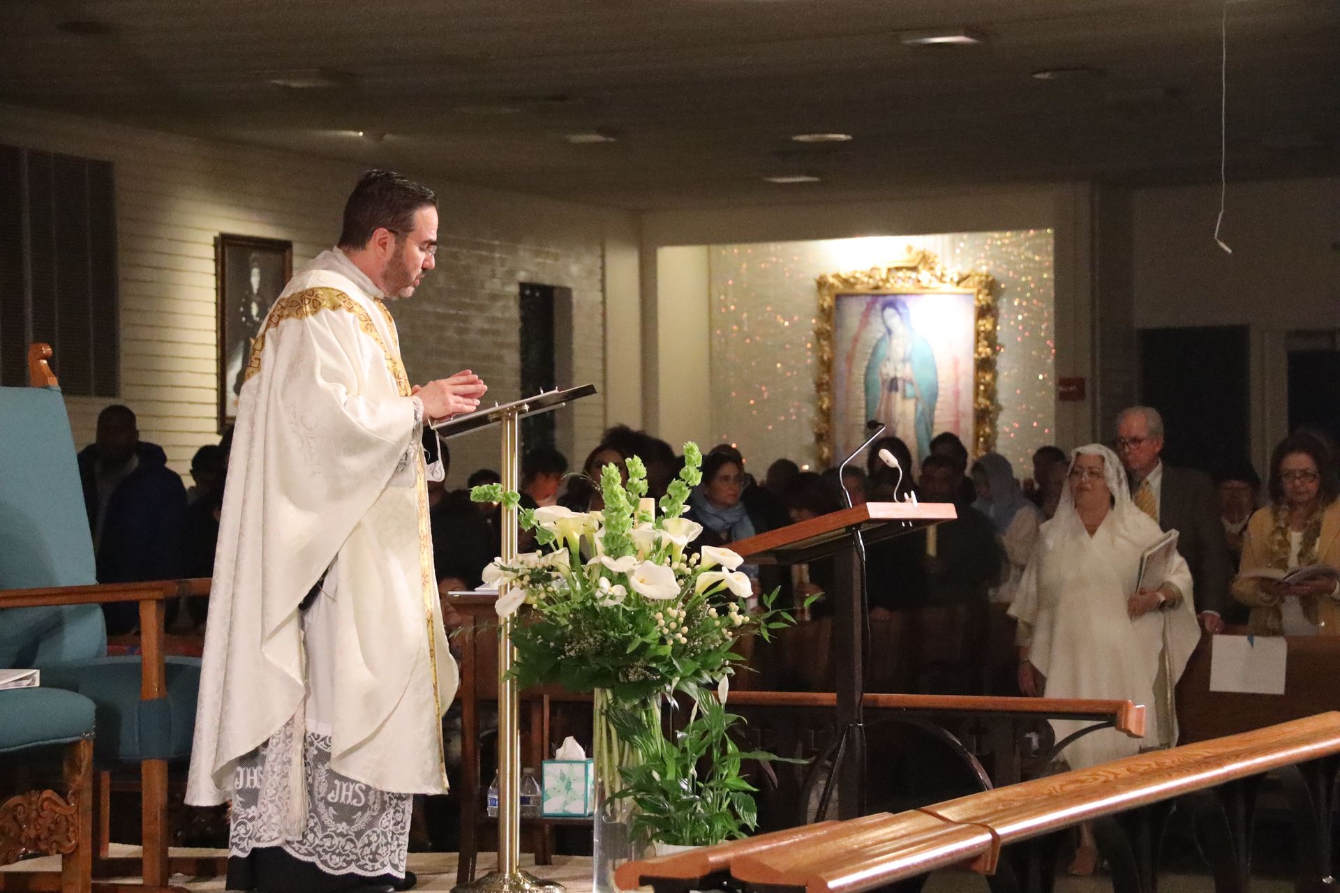 Priest in white vestments speaking at a church altar during a service