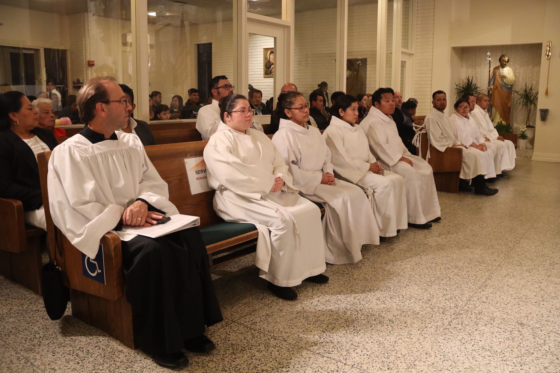 People seated in a church pew during a religious service, wearing white robes and facing forward.