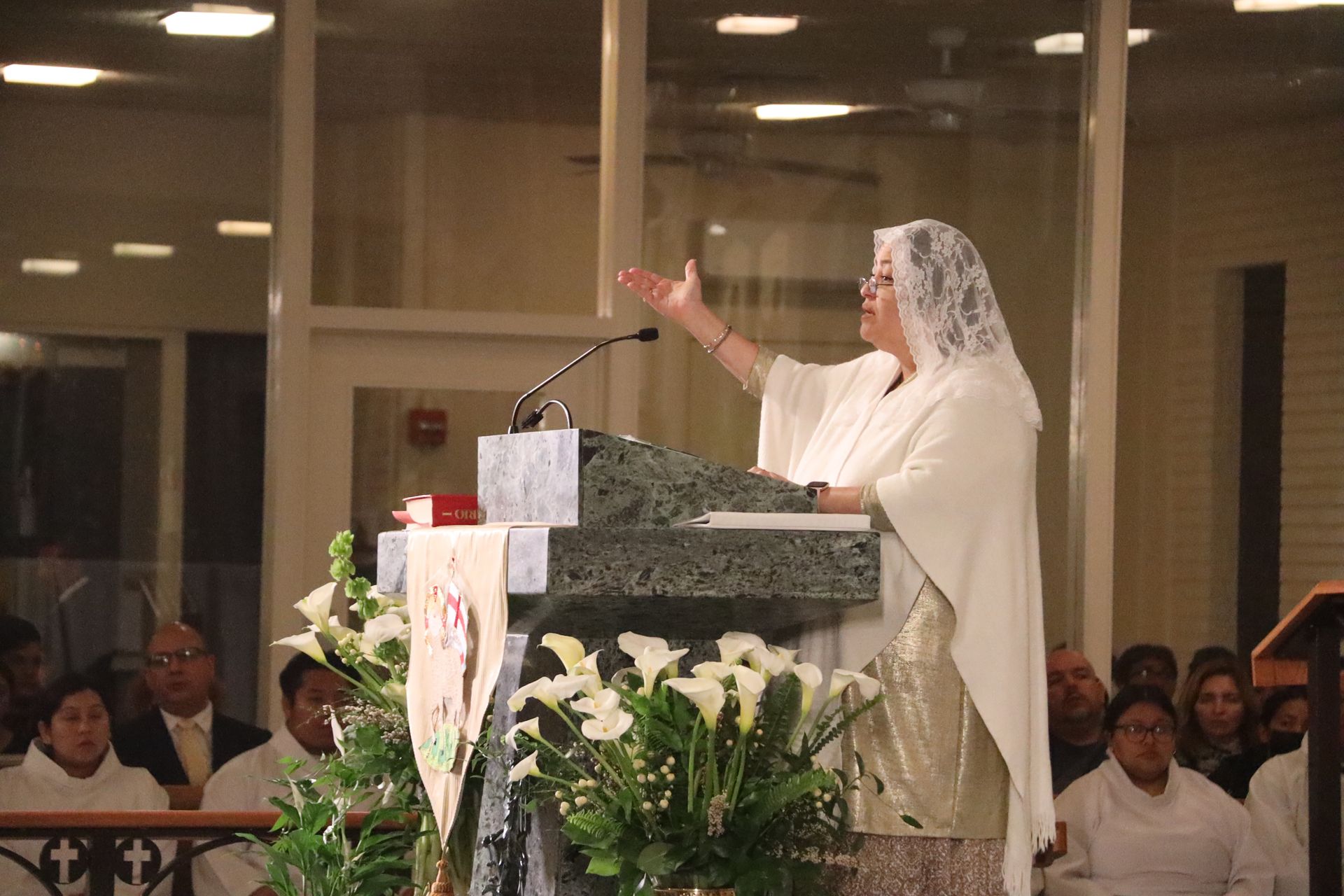 Woman in white speaking at a podium in a church, raising her hand to address the congregation.