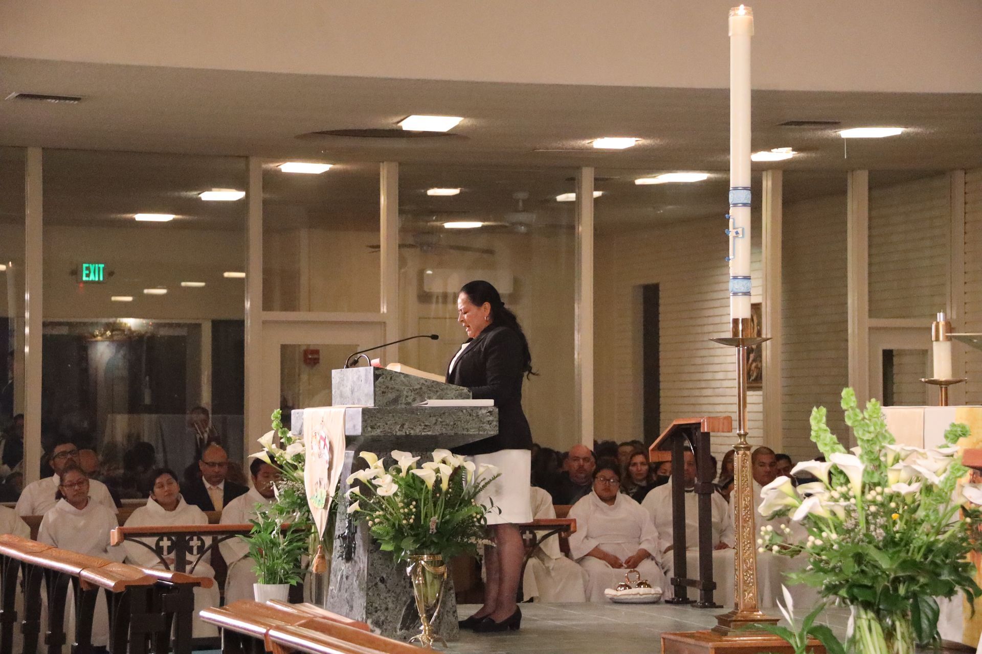 Speaker at podium in a church, addressing a seated congregation with floral arrangements in front.