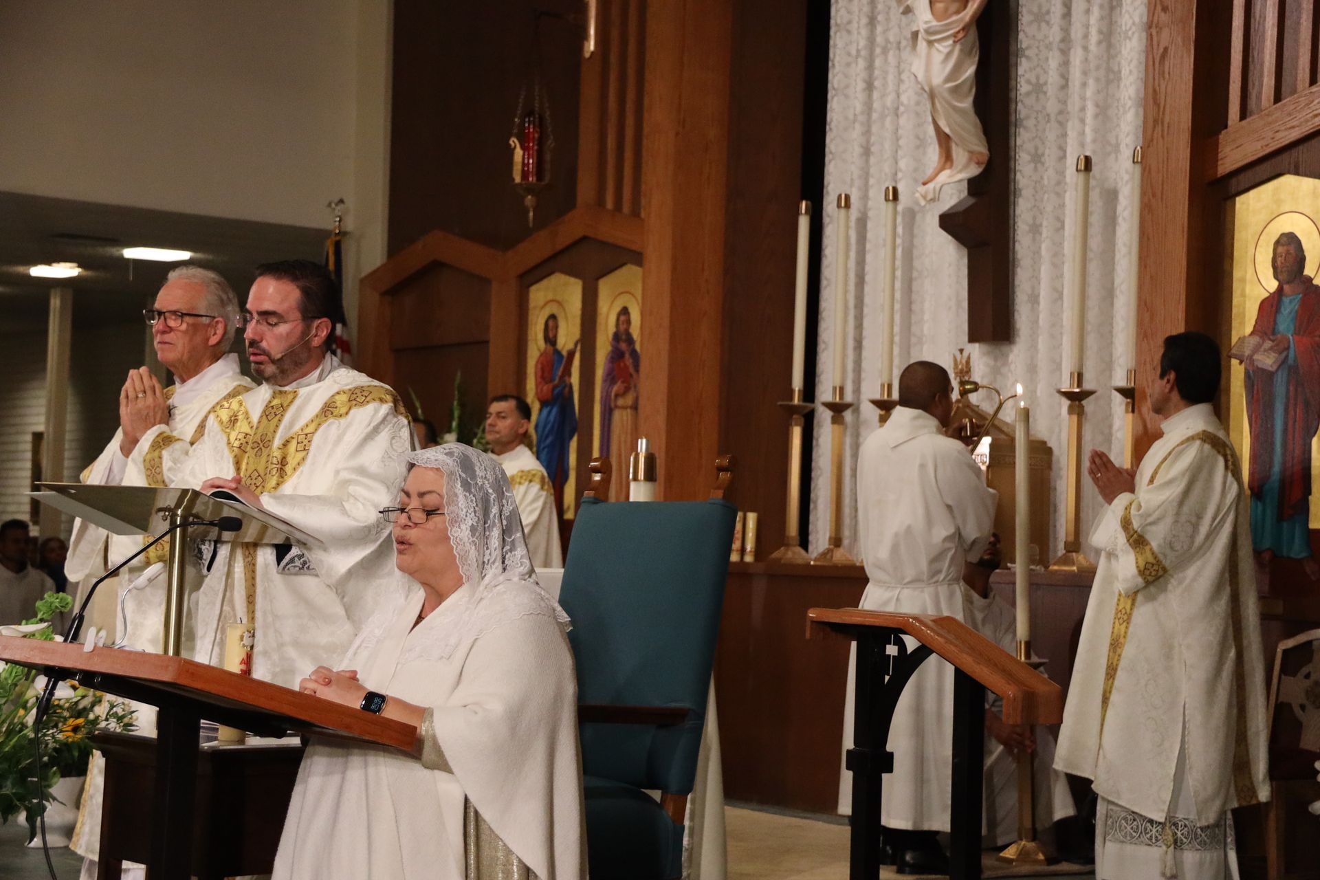 Clergy in white robes conduct a church service at the altar with a seated woman reading nearby.