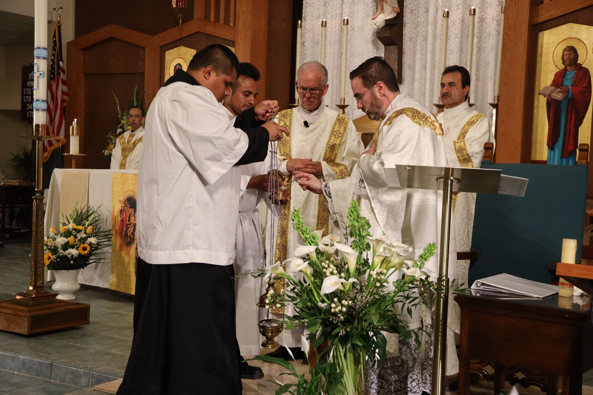 Priests in white vestments celebrate Mass at an altar with flowers in a church.