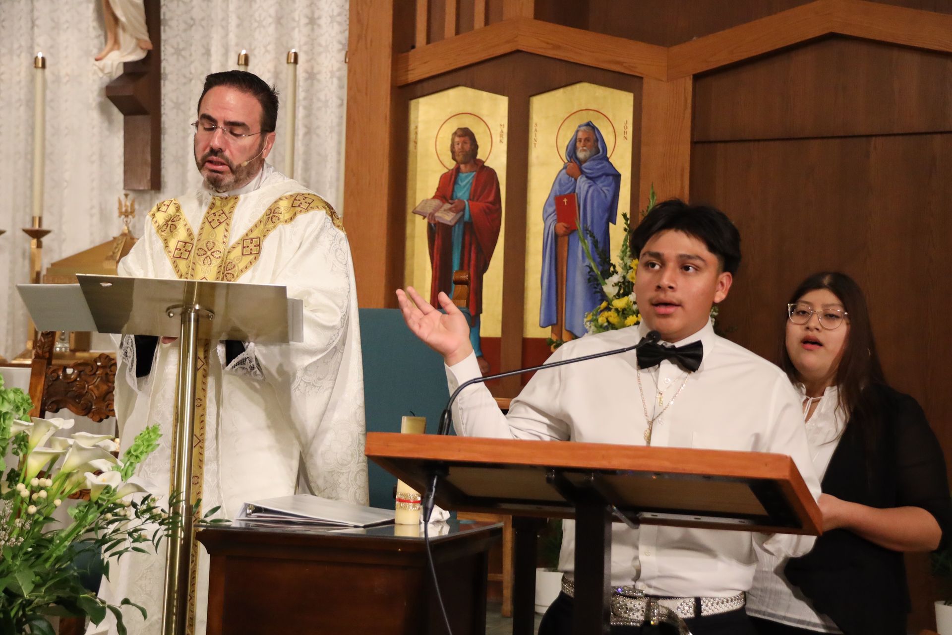Clergy in white robes at a church lectern with a boy and woman singing or speaking beside him