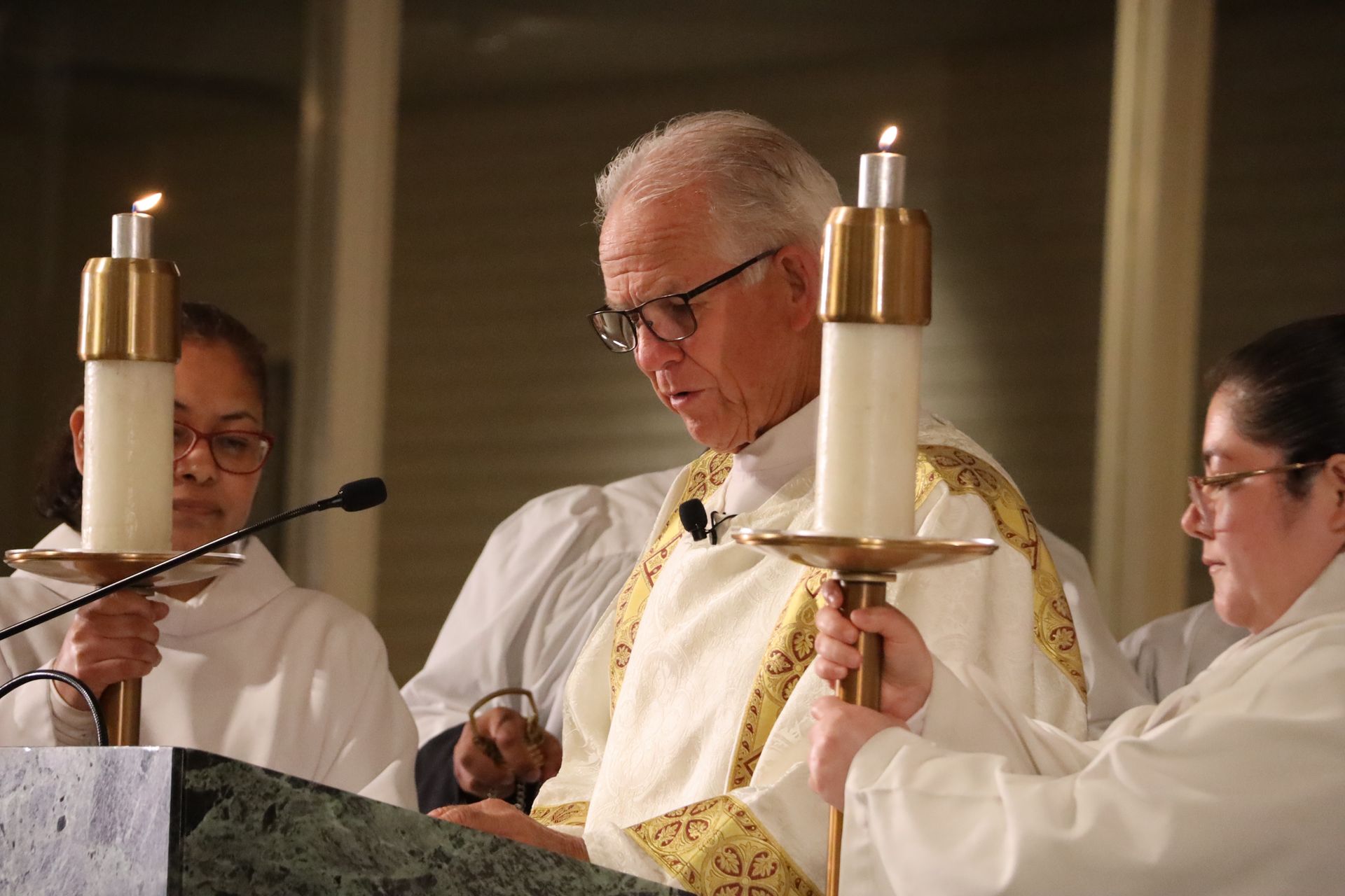 Clergy in white and gold robes stand at an altar during a church service, holding lit candles.