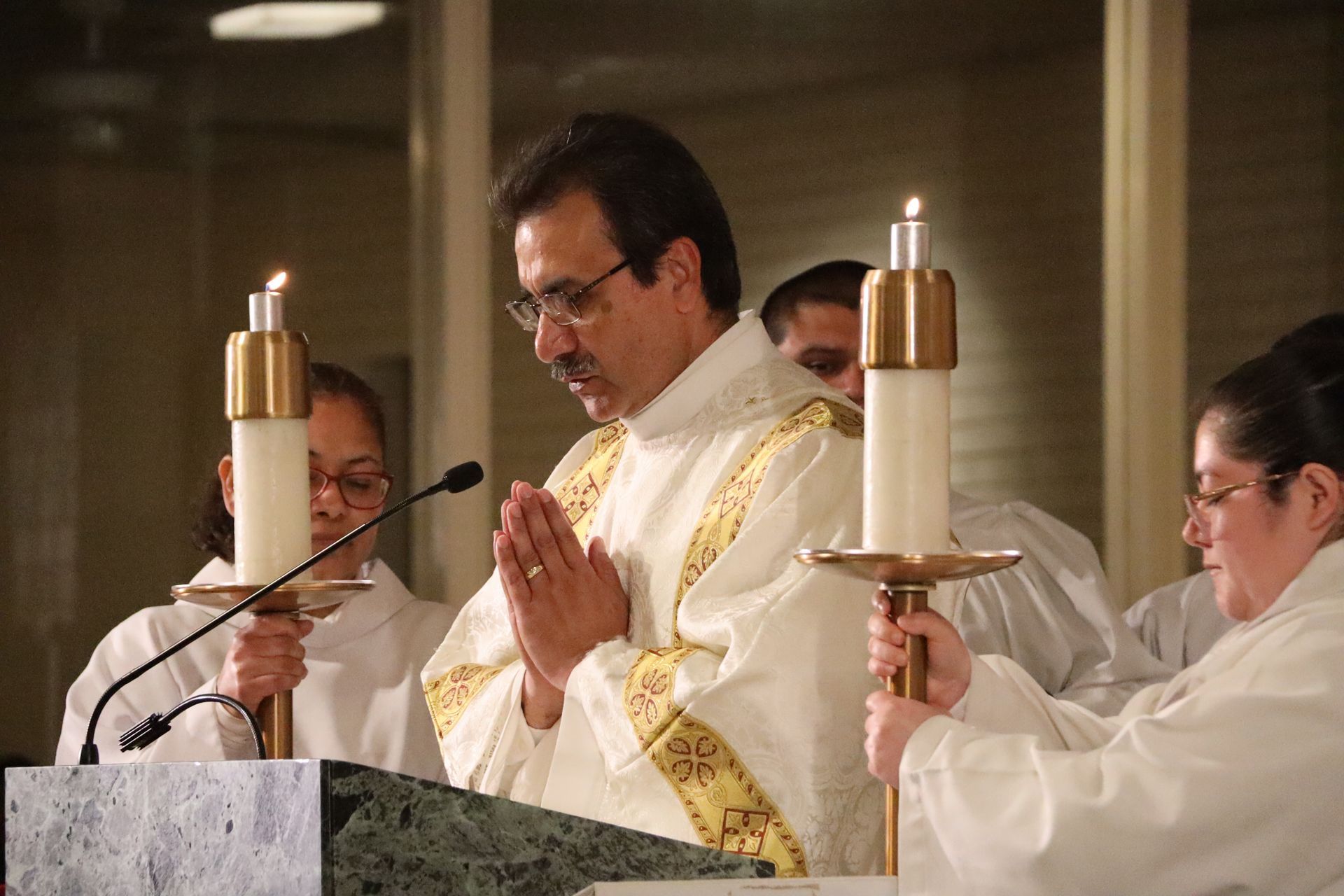 Priest celebrating Mass at an altar, flanked by assistants holding candles, in a church interior