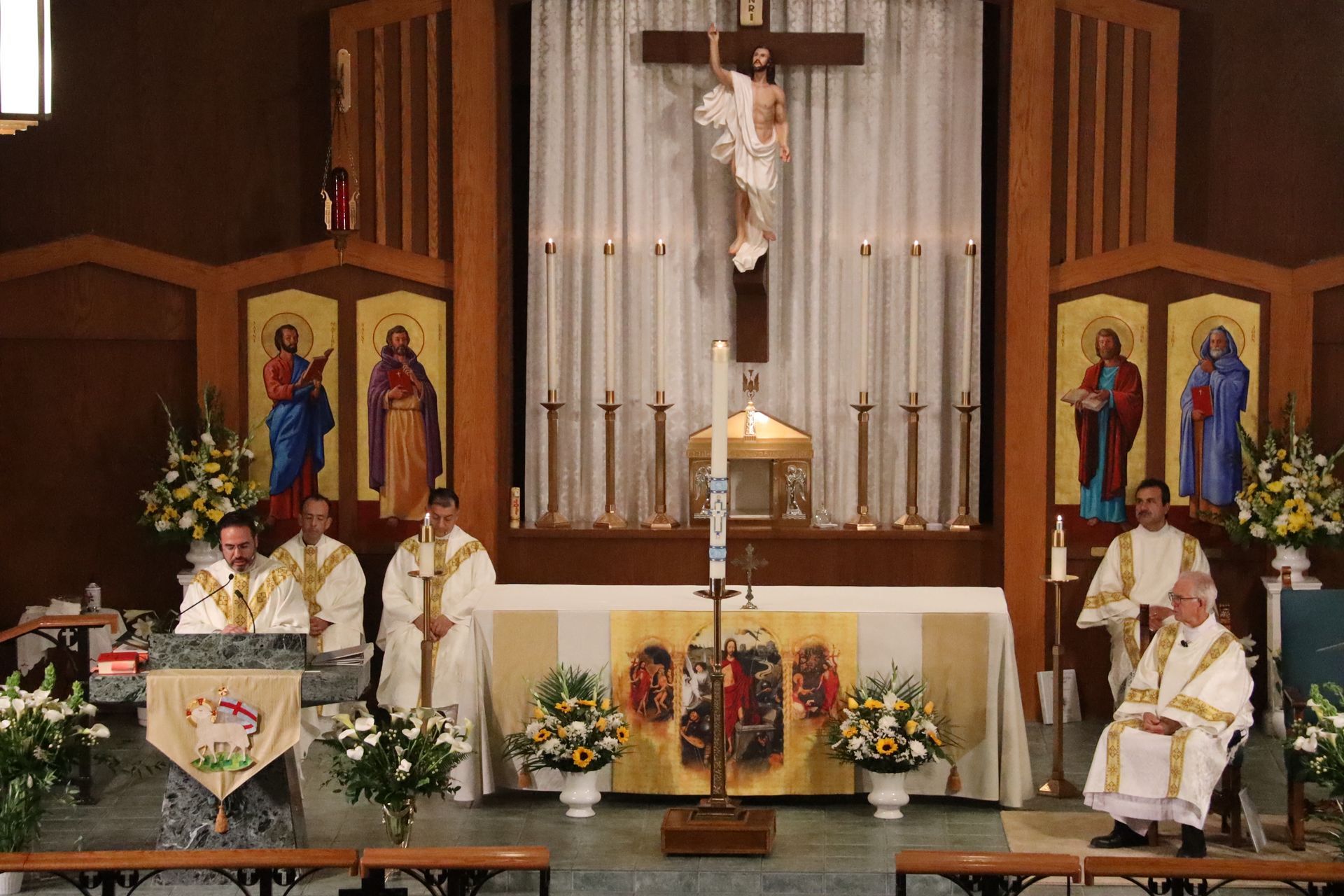 Catholic church altar during Mass, with priests in white vestments, candles, flowers, and a crucifix centered above.