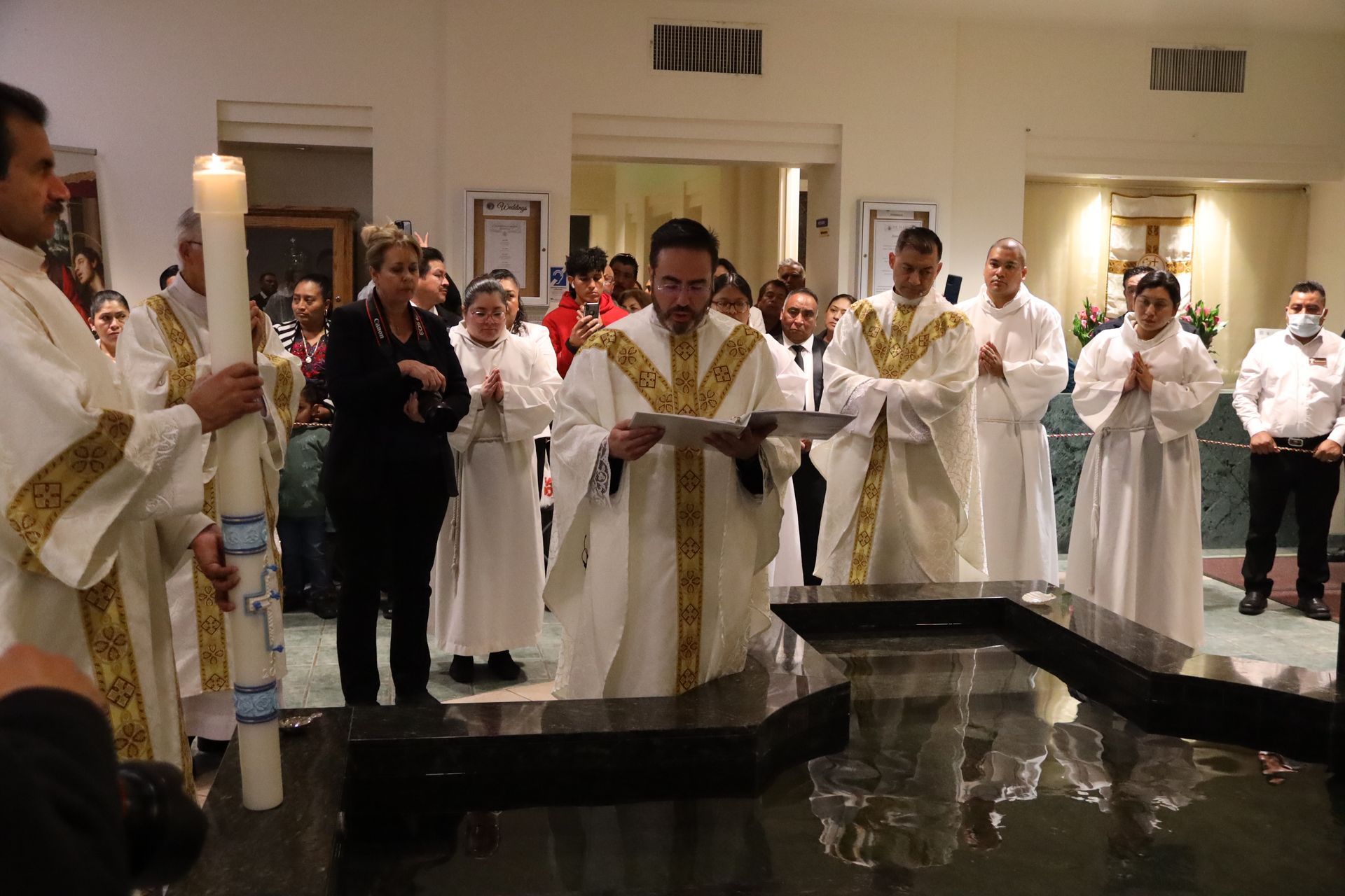 People in white robes and a priest gathered around a baptismal font in a church hall.