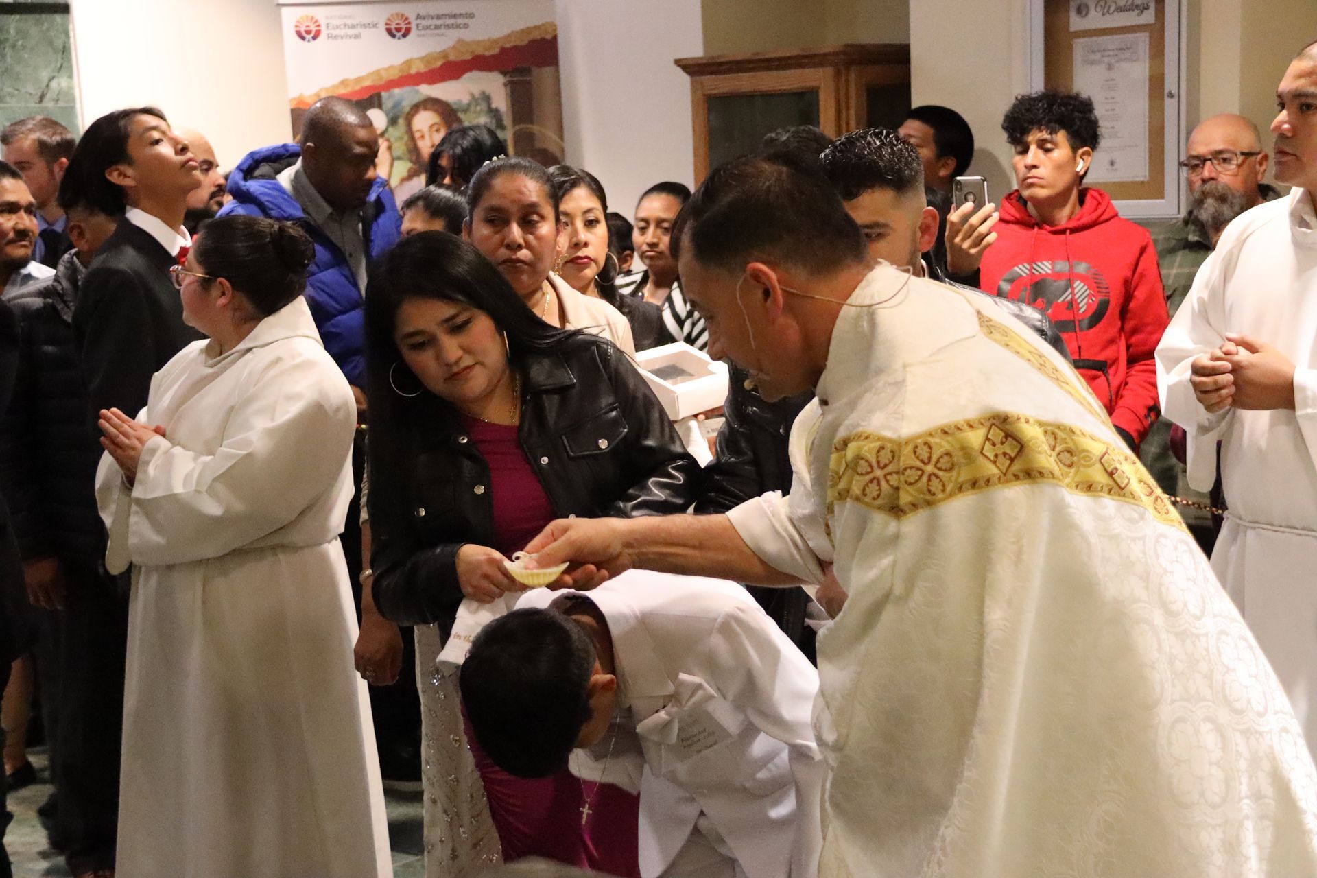 Priest blessing a child during a crowded church ceremony, with onlookers nearby
