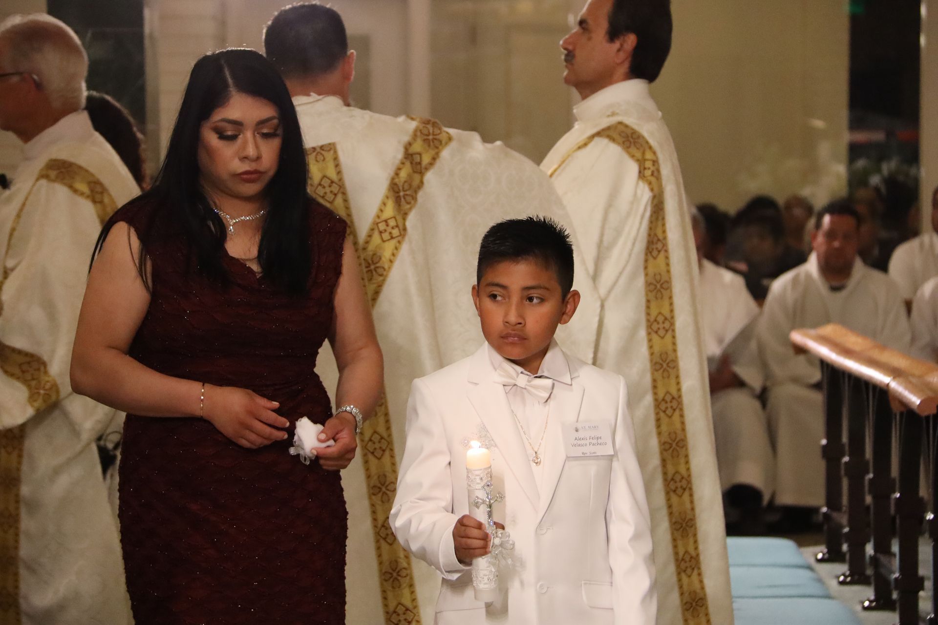 A woman and a boy in white stand in a church aisle during a ceremony, each holding a candle.