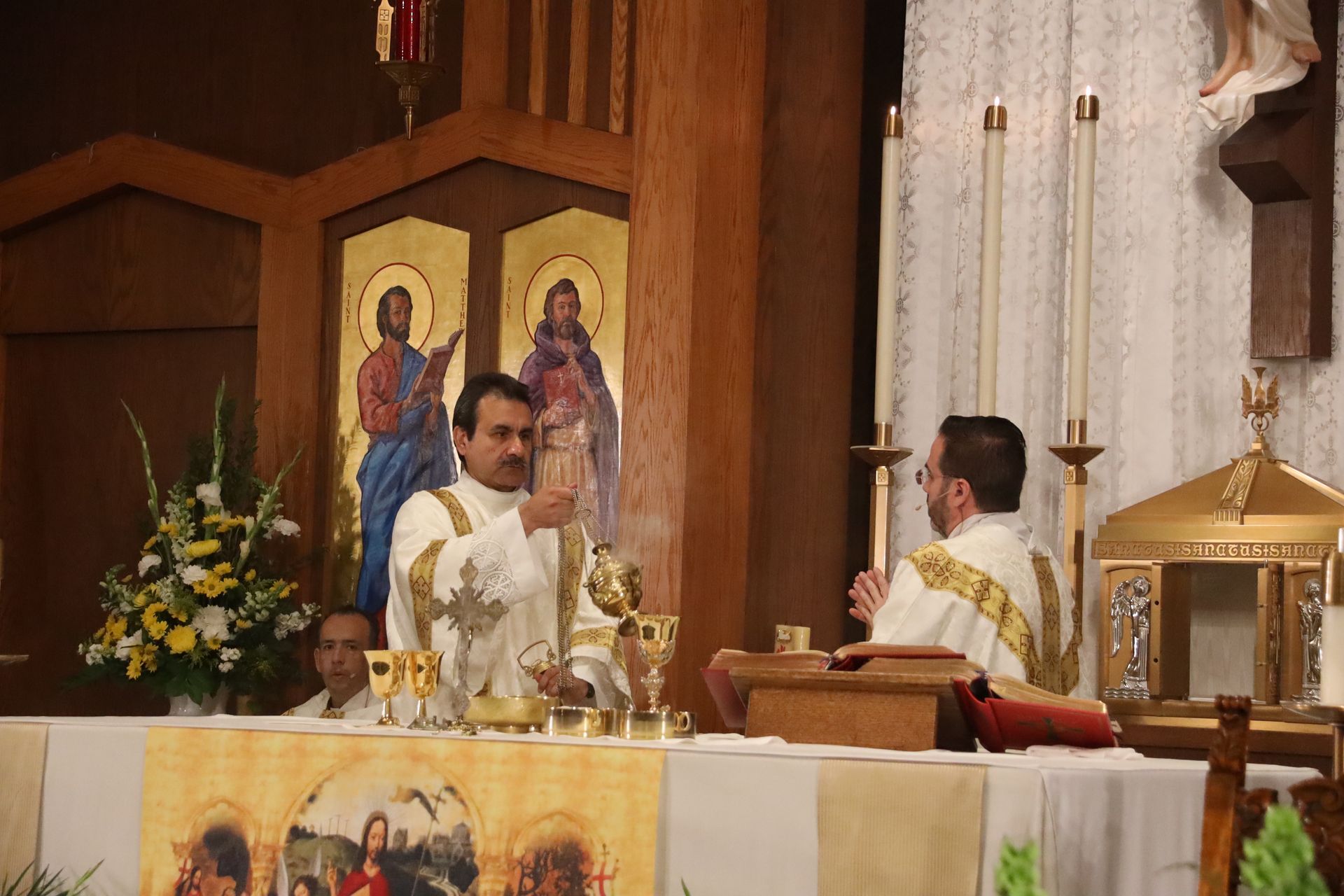 Two priests celebrate Mass at an altar in a church, with a gold tabernacle and religious icons behind them