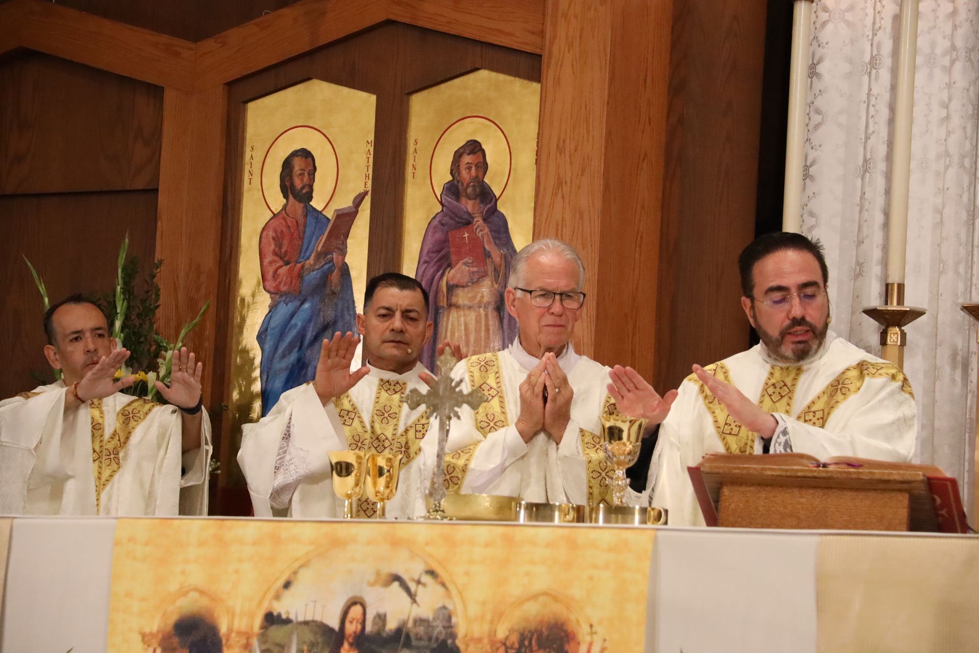 Four clergy in white robes stand at an altar during a church service, with religious icons behind them.