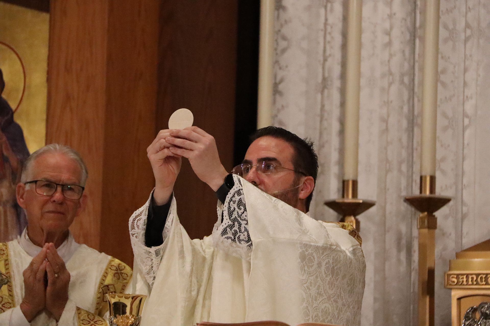 Priest holding up a communion wafer during Mass, with another priest praying beside him
