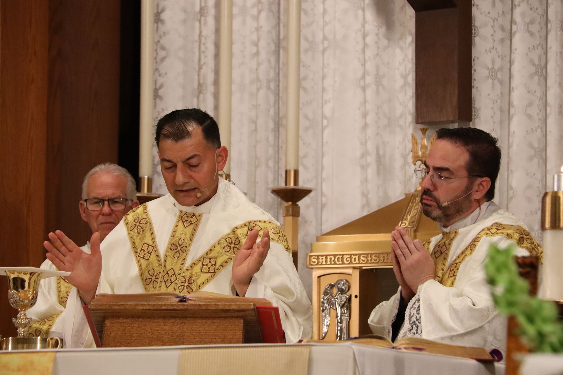 Three clergy in white and gold vestments praying at an altar during Mass