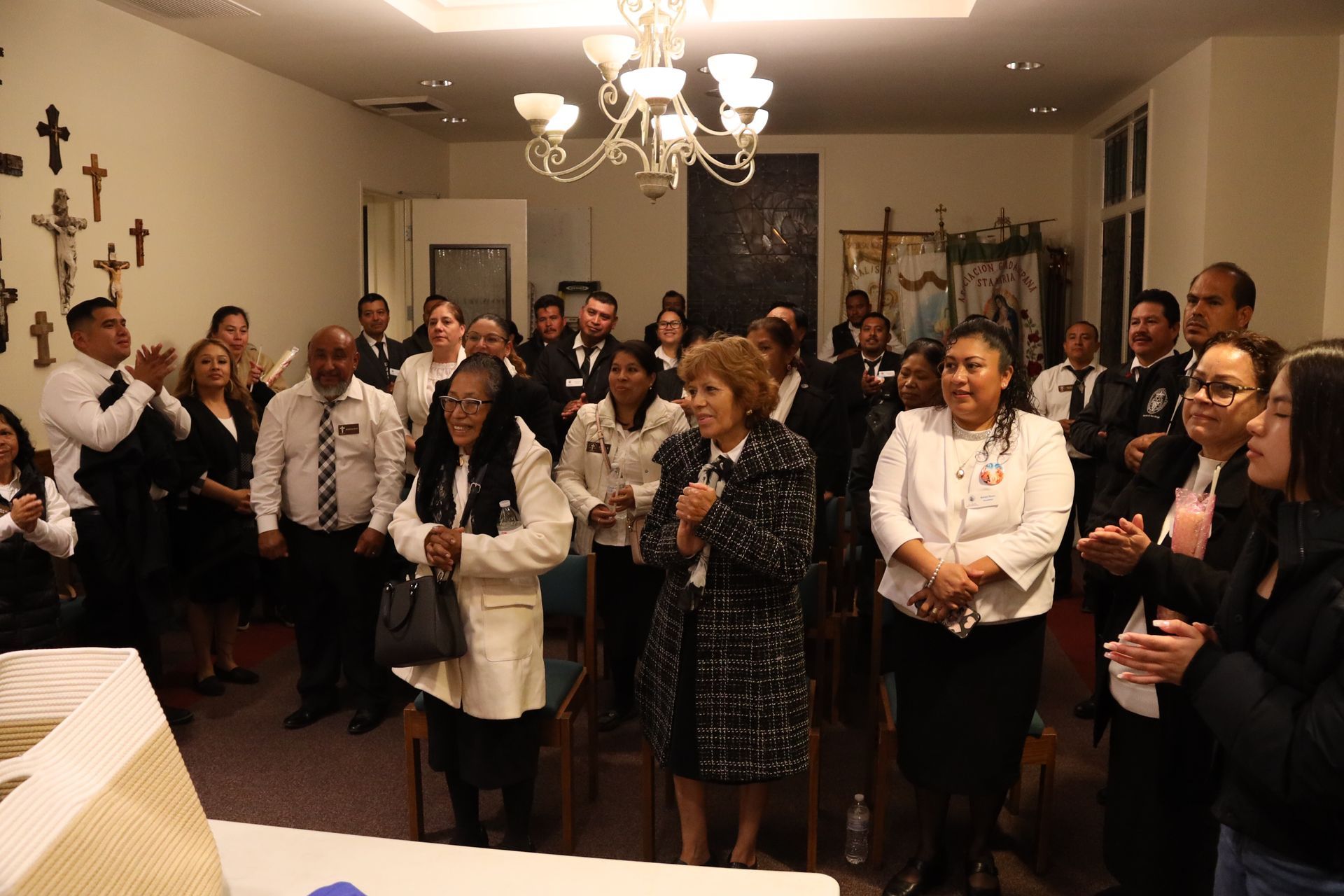 People gathered indoors, clapping in a warmly lit room with wall crosses and a chandelier.