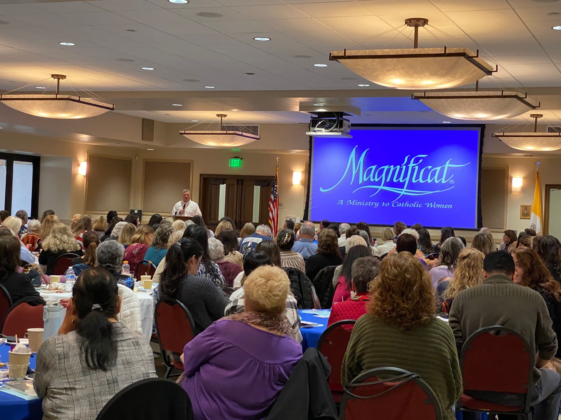 Crowded conference room with seated audience facing a large screen showing “Magnificat” in blue.