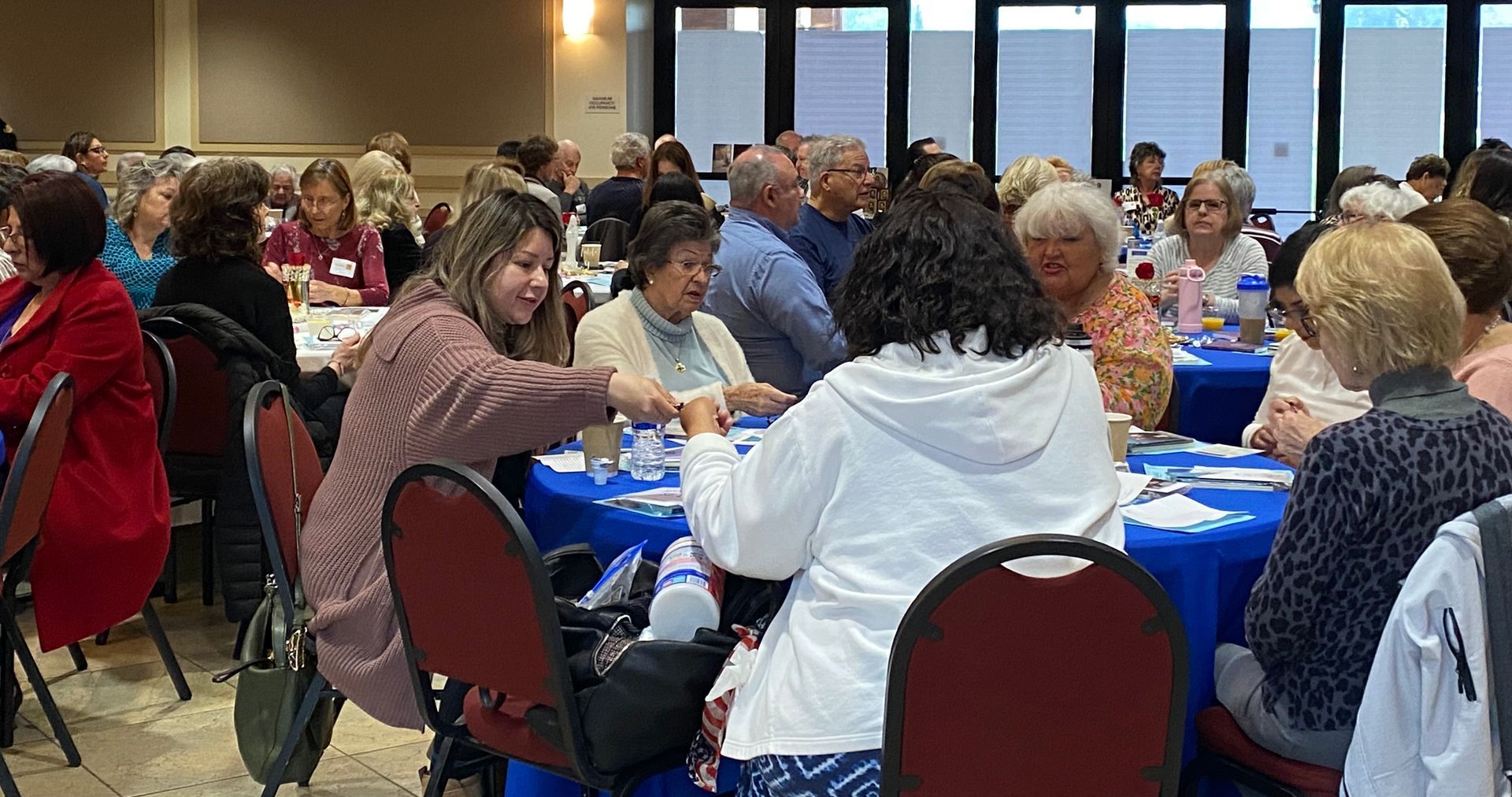 People seated at round tables in a banquet hall, talking and eating during a community event.