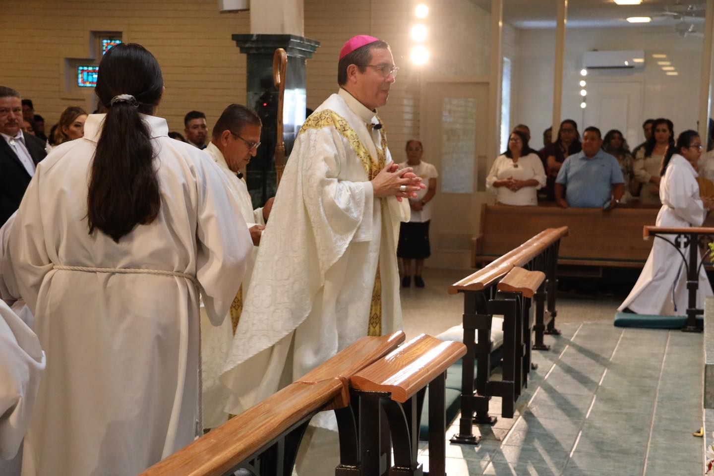 Priest in white vestments leads a church service with attendants standing beside pews inside a chapel