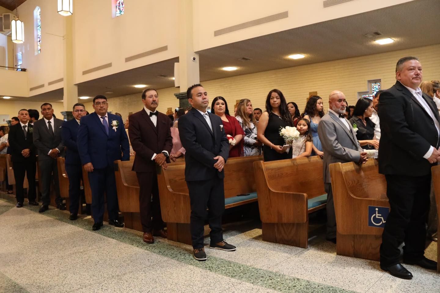 People standing in church pews during a service, with one man in a wheelchair near the front.