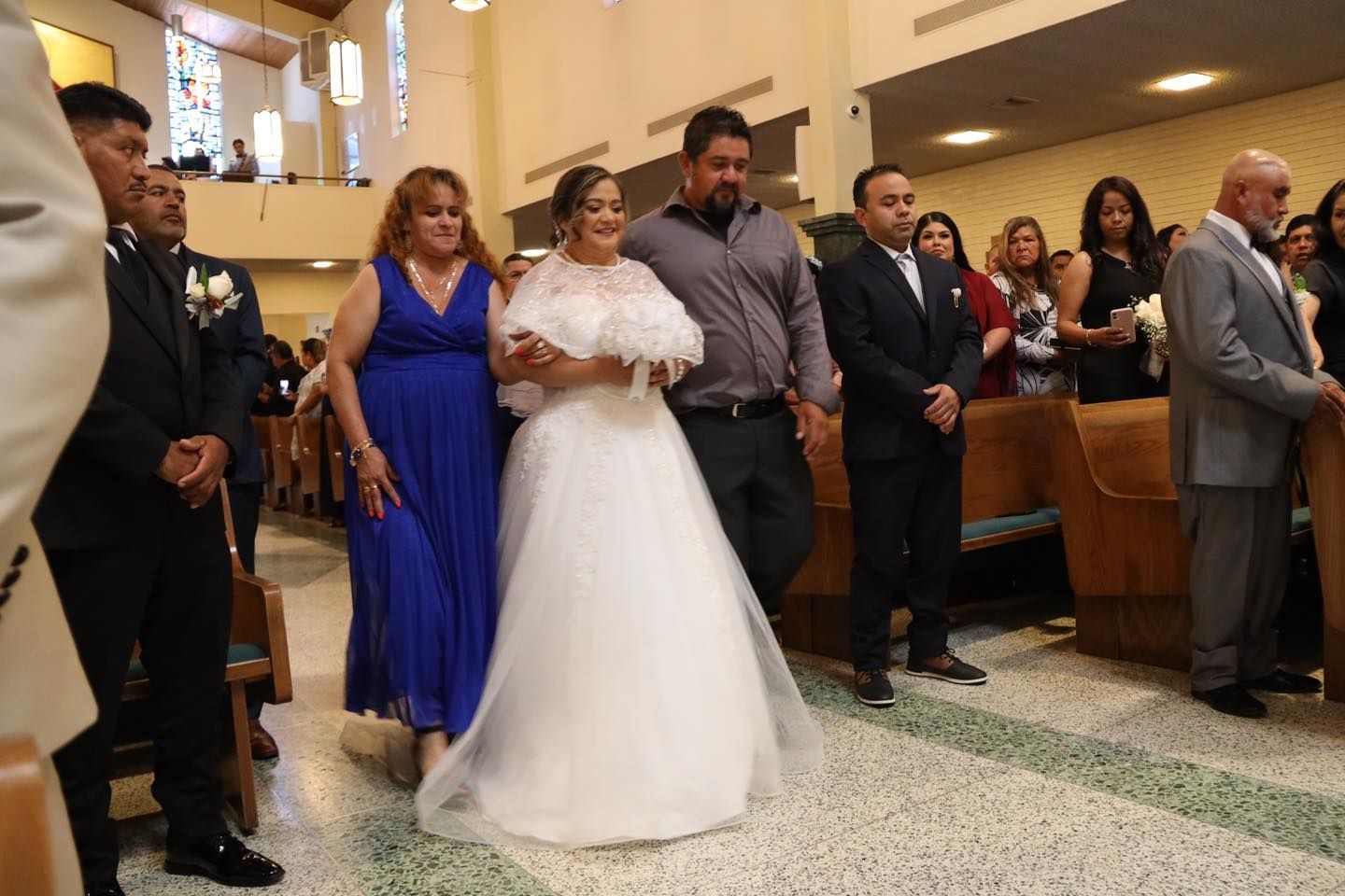 Bride in white gown walks down church aisle with attendants in blue dresses and a man in a gray shirt
