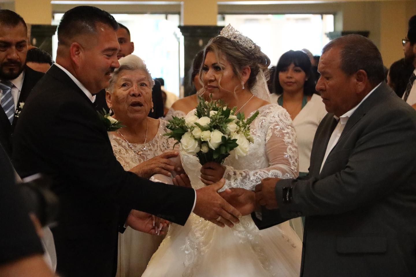 Bride being escorted down the aisle, holding a white bouquet, surrounded by family in a church.