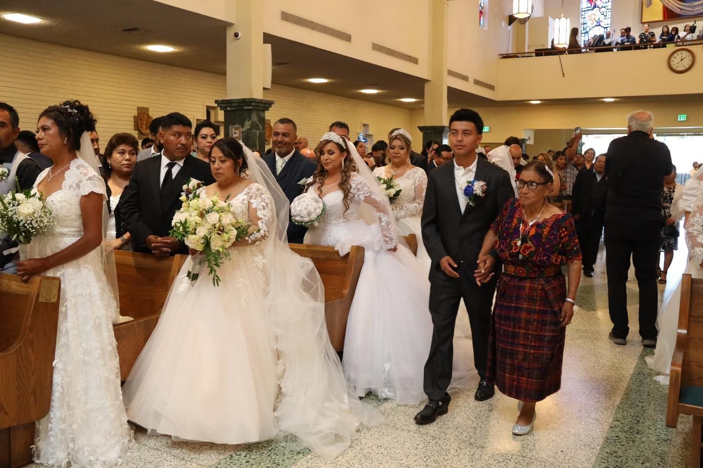 Bridal party walking indoors at a wedding reception, women in white gowns carrying bouquets, one man in a dark suit.