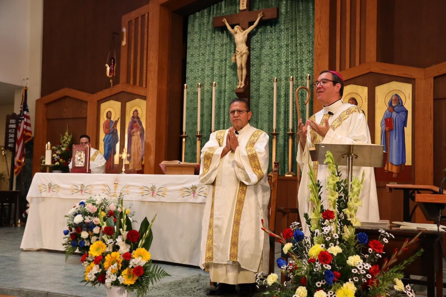 Catholic altar scene with two priests at the lectern, crucifix, candles, and floral arrangements.