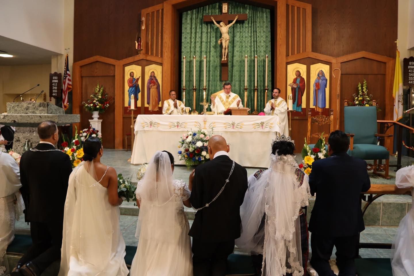 Children in white veils and dark suits face an altar during a church ceremony under a crucifix.