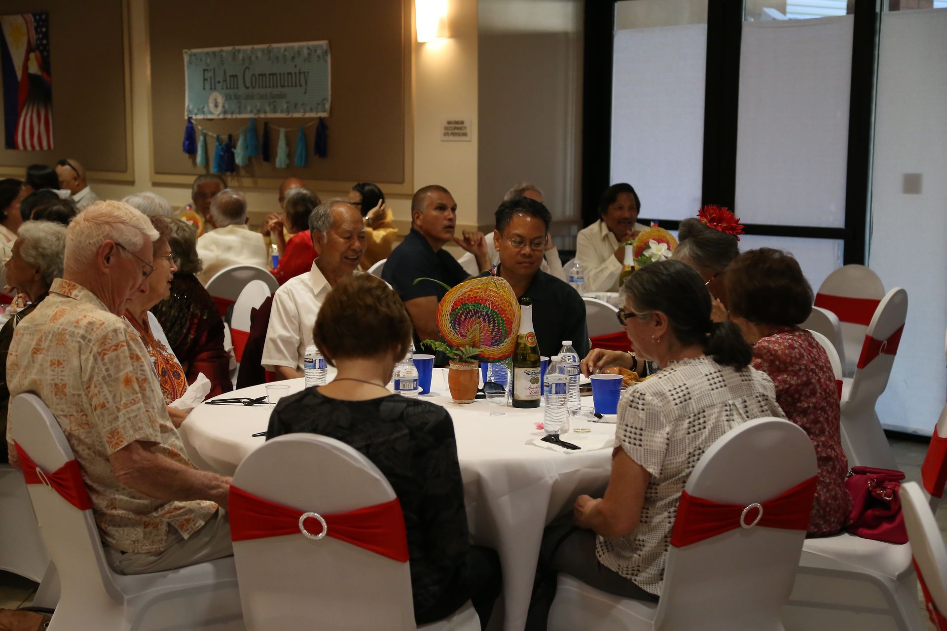 People seated at round tables in a banquet hall, listening during a community gathering.