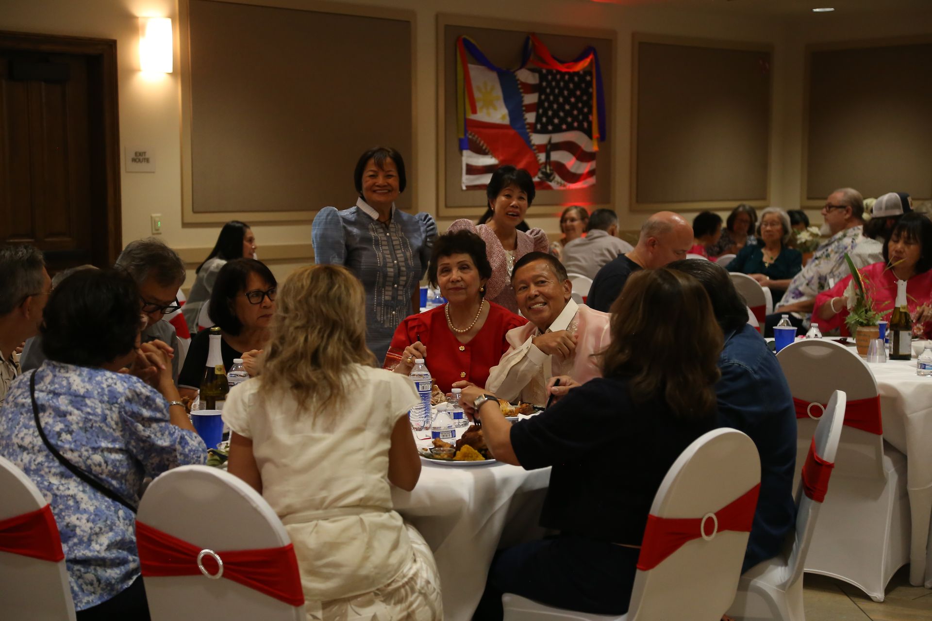 People seated around banquet tables in a decorated conference room, listening to a speaker near the back.
