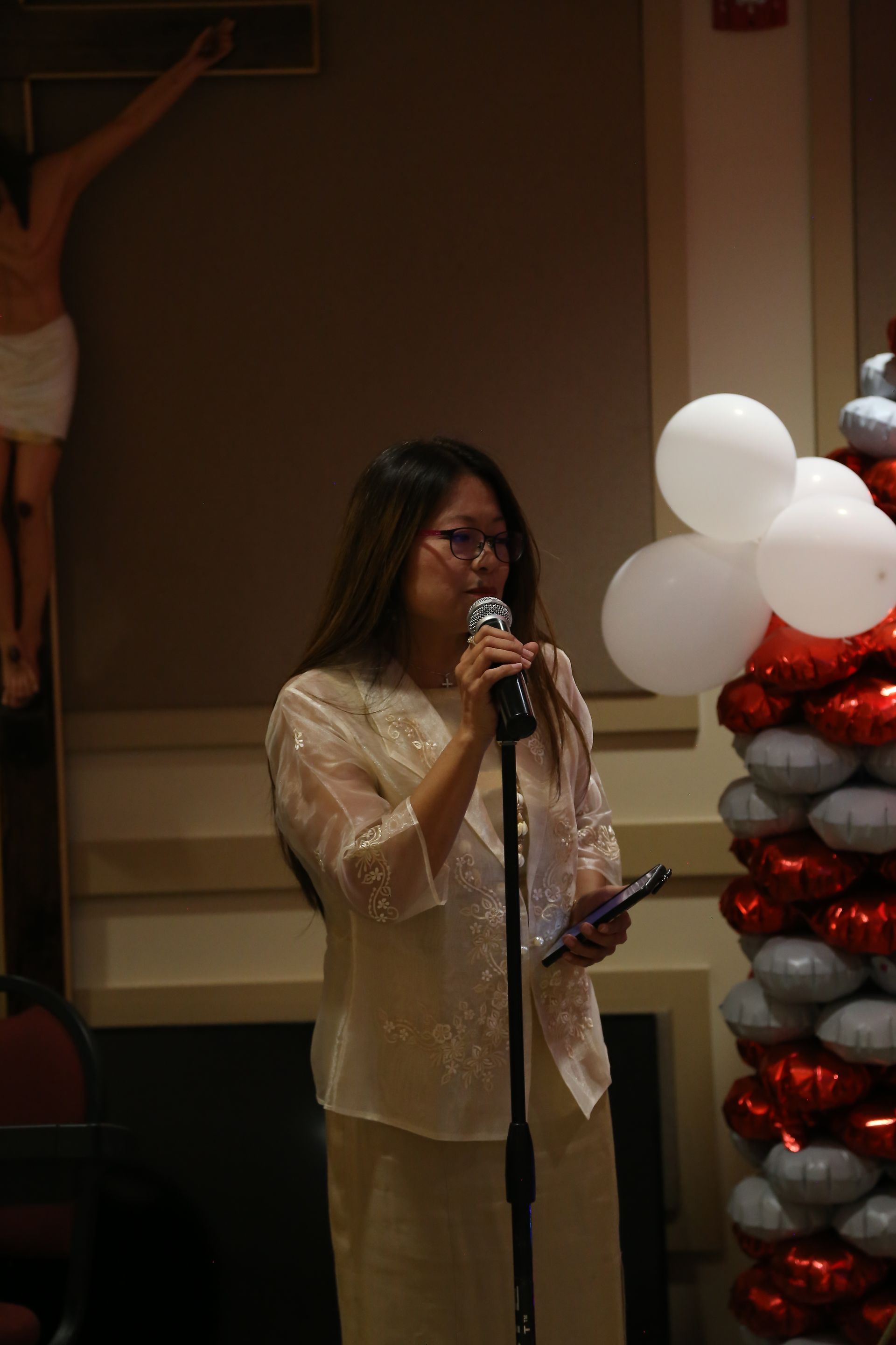 Woman speaking at a microphone beside red and white balloon decorations at an indoor event