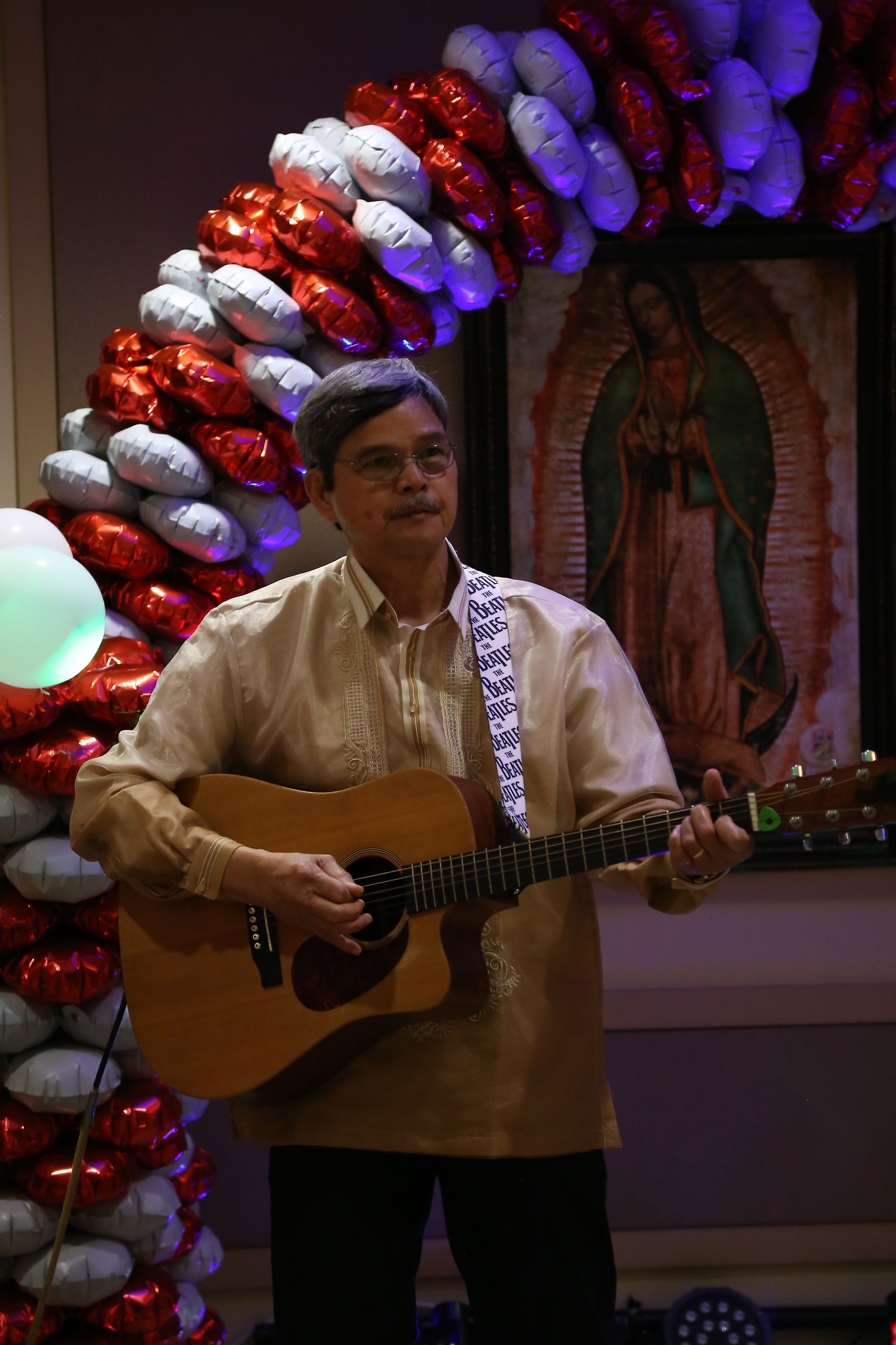 Man playing acoustic guitar beside a colorful balloon arch and framed religious image