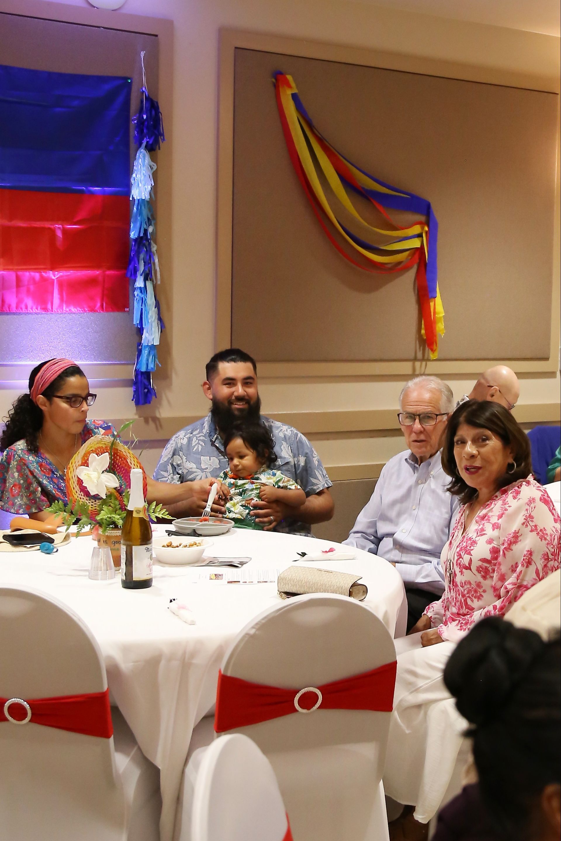 People seated at a banquet table in a decorated room with colorful wall streamers and balloons