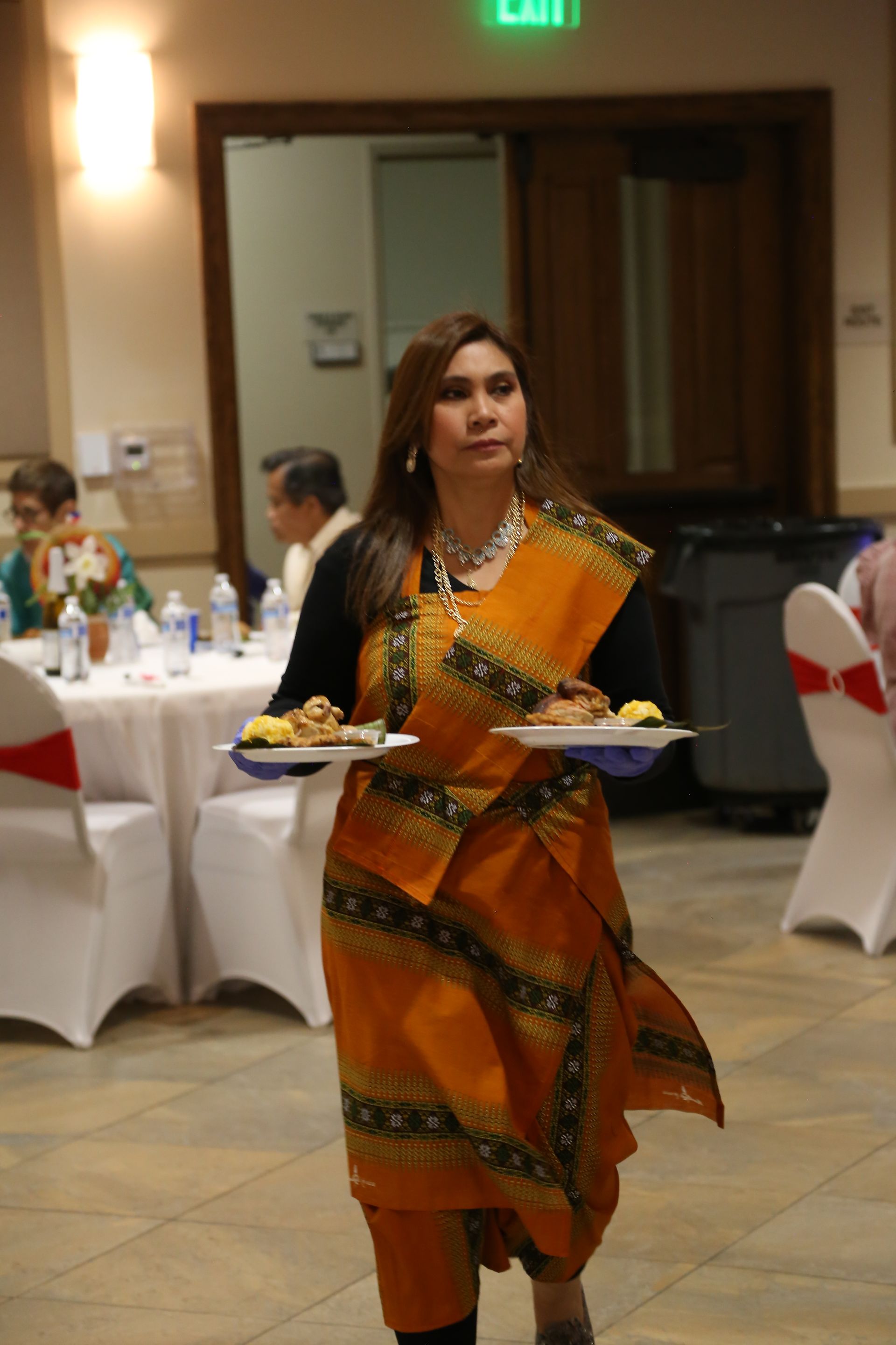 Woman in orange patterned dress carrying two plates at a banquet hall event