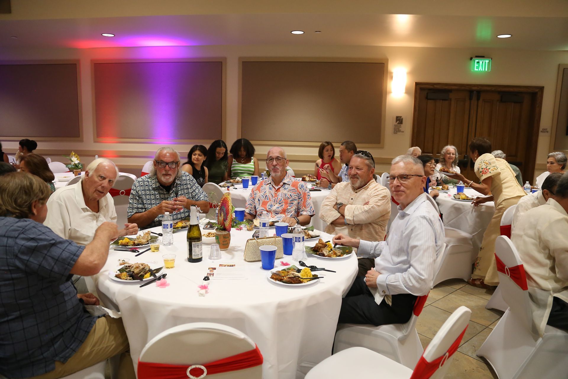 Guests seated at round tables in a banquet hall, eating and chatting under purple accent lighting