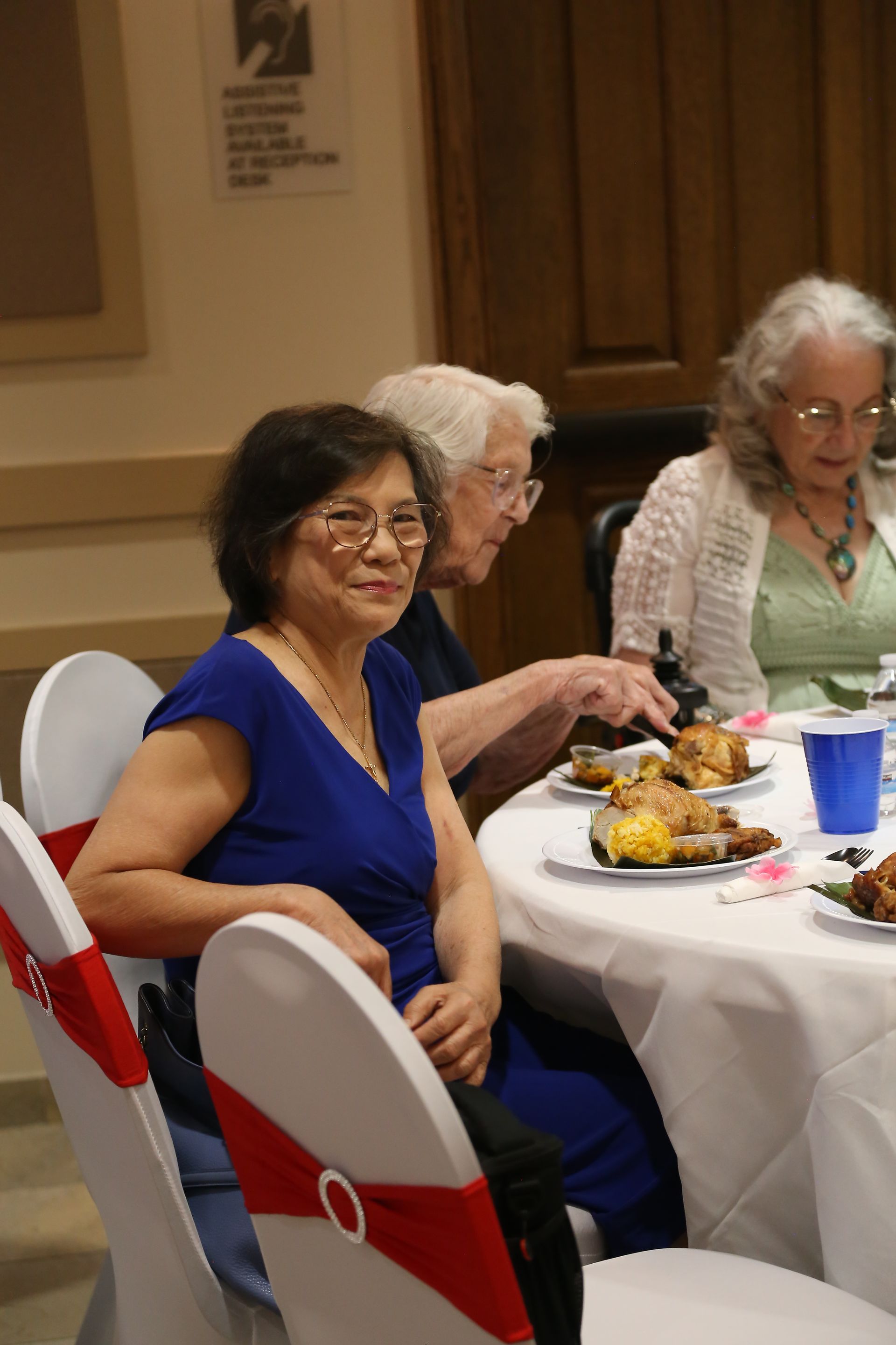 People seated at a banquet table, one woman in a blue dress reaching for food.