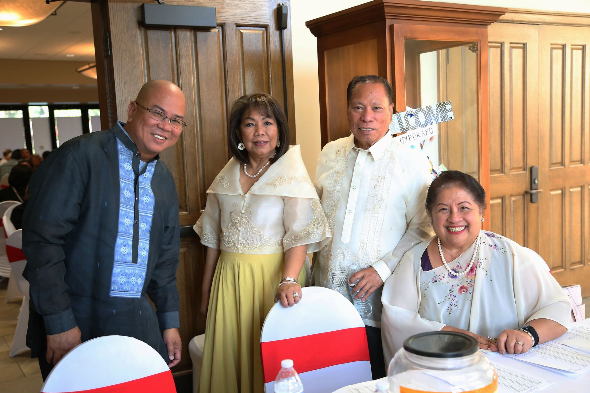 Four people posing indoors at a table, smiling and wearing formal attire with red-and-white chair covers.