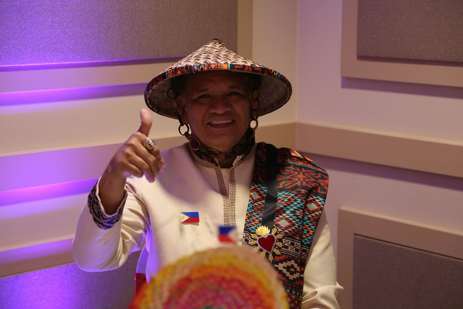Person smiling and giving a thumbs-up, wearing a patterned hat and traditional-style clothing indoors.